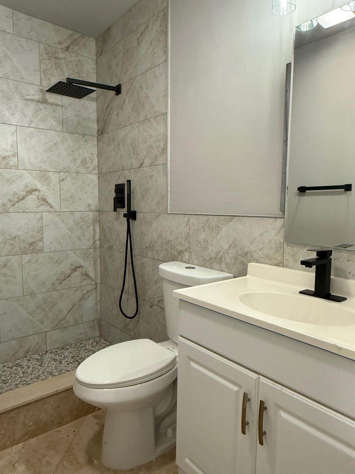 Bathroom with a marble-look tiled shower, black fixtures, pebble floor, toilet, and white vanity.