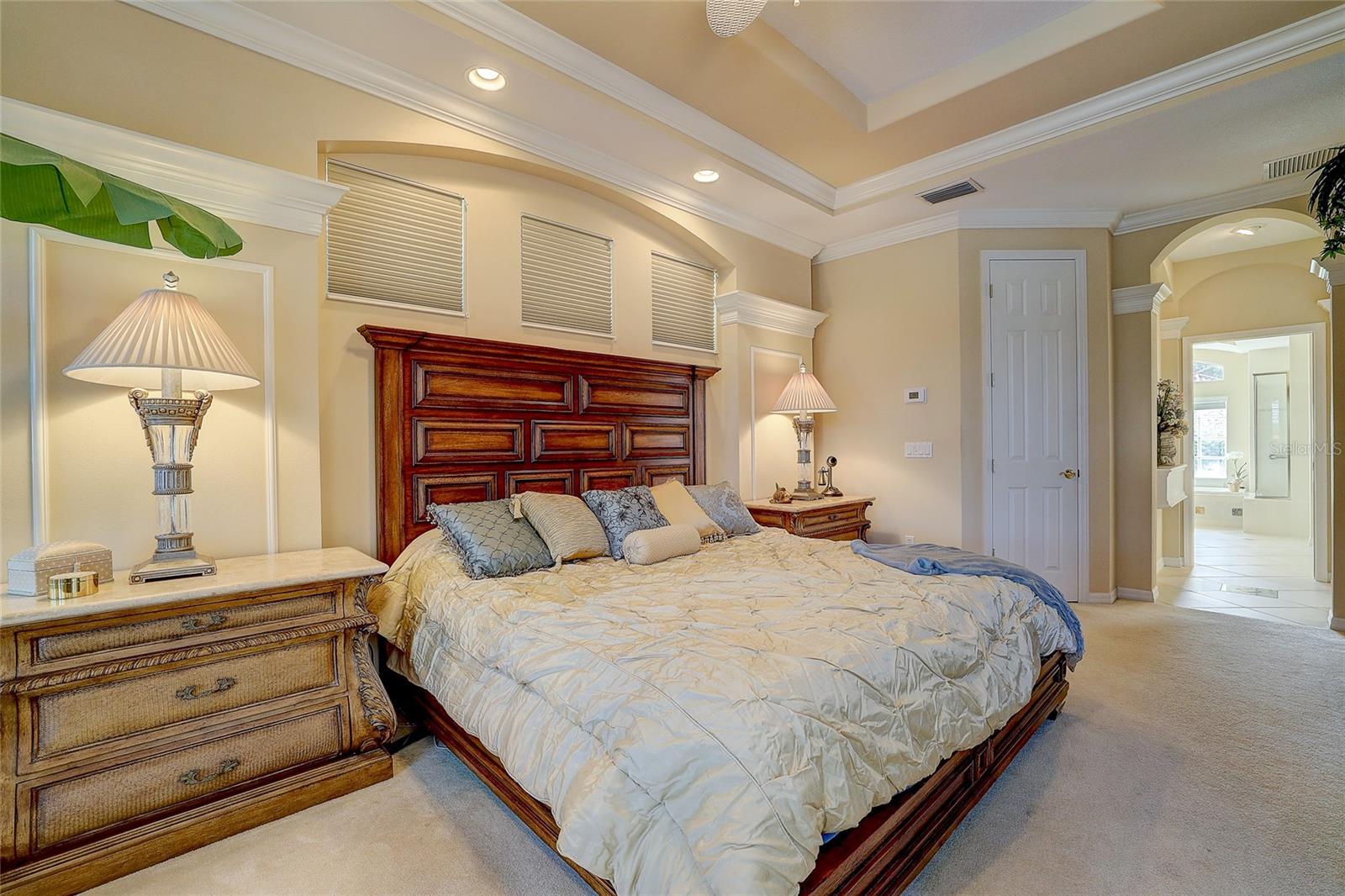 Primary bedroom with an ornate dark wood headboard, tray ceiling, and doorway to a bright bathroom.