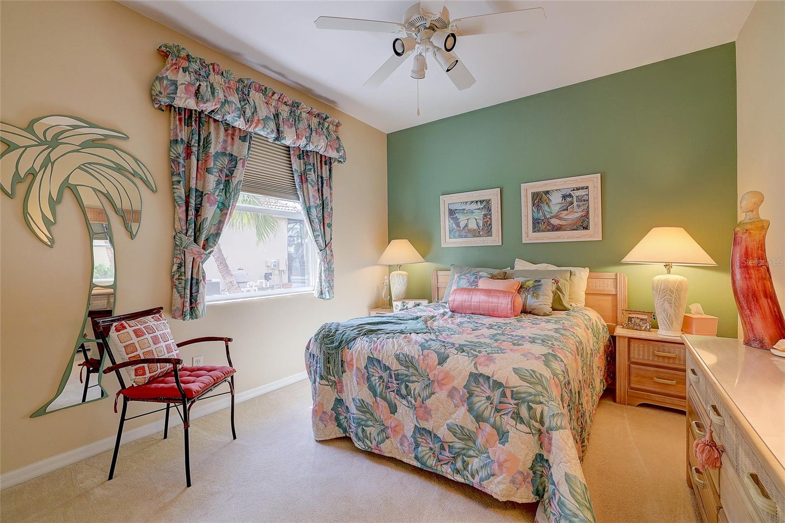 Bedroom with a floral bedspread, green accent wall, tropical curtains, and palm-tree-shaped mirror.