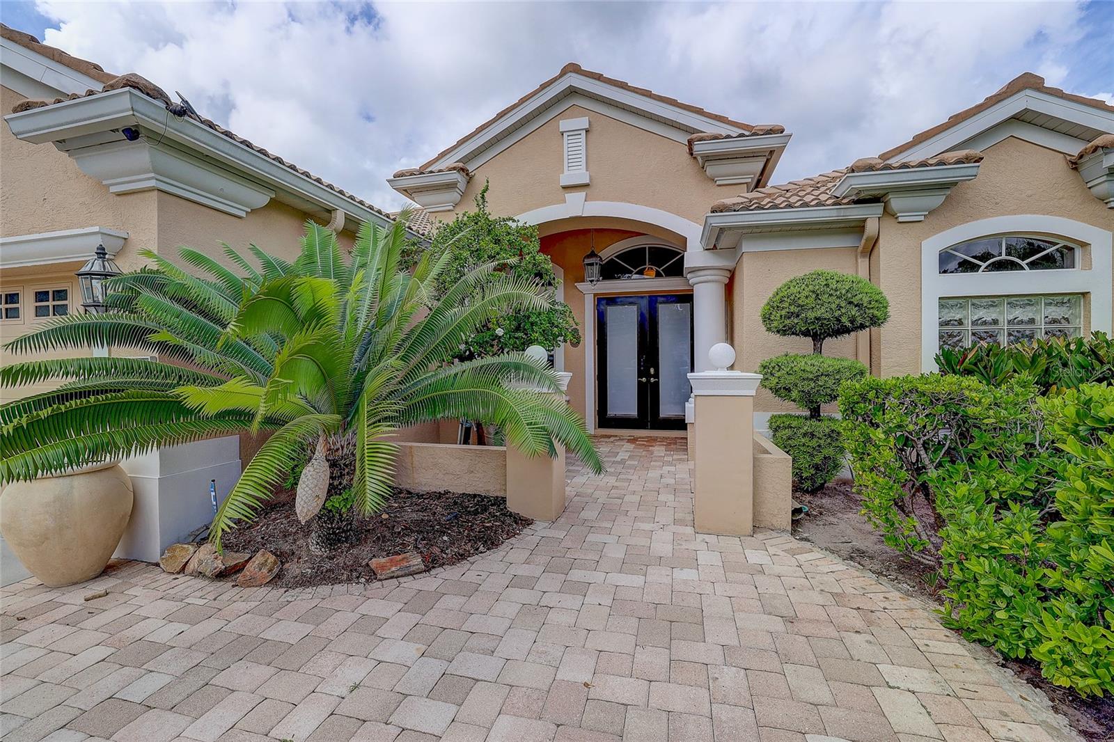 Beige stucco home entrance with arched entry, paver walkway, palms, and manicured shrubs.
