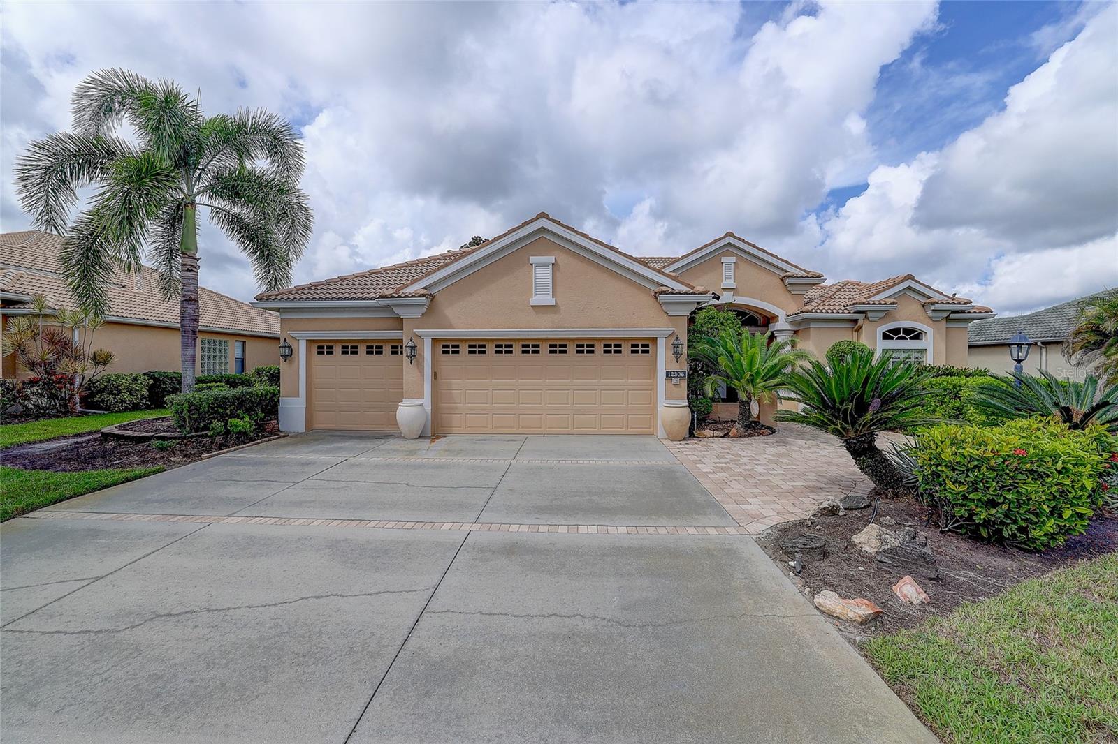 Tan stucco home with a three-car garage, wide driveway, and palm trees by the entrance.