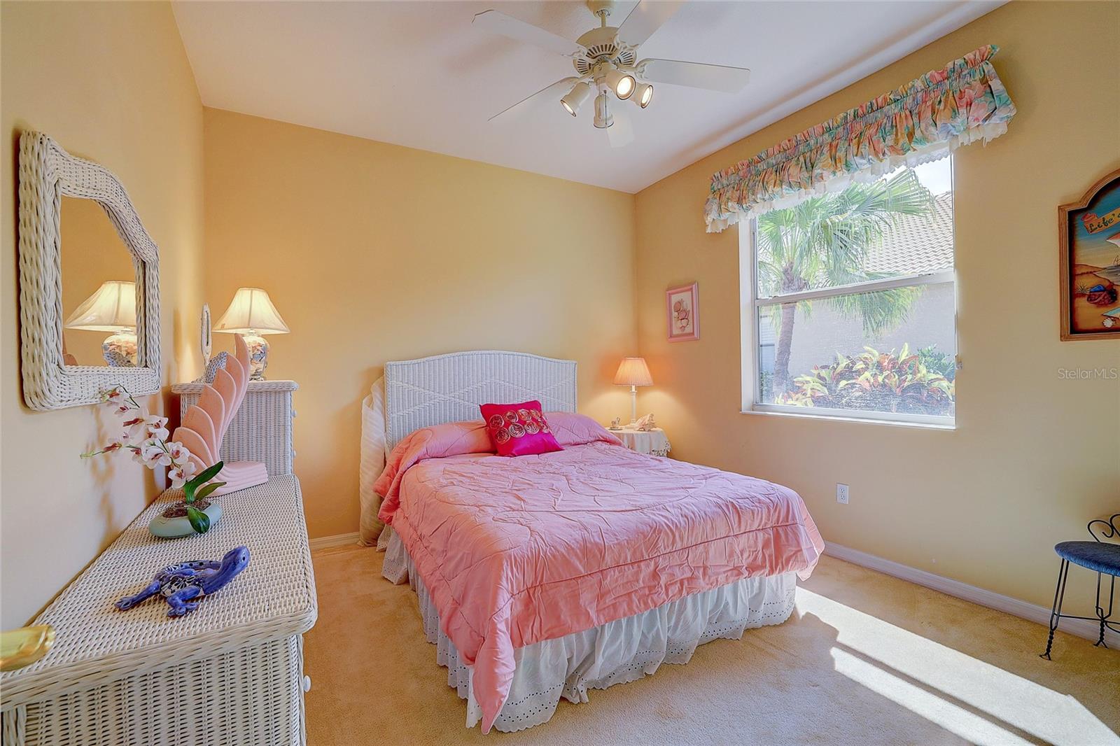 Bedroom with a pink quilted bed, white wicker furniture, and sunlight streaming through a window.