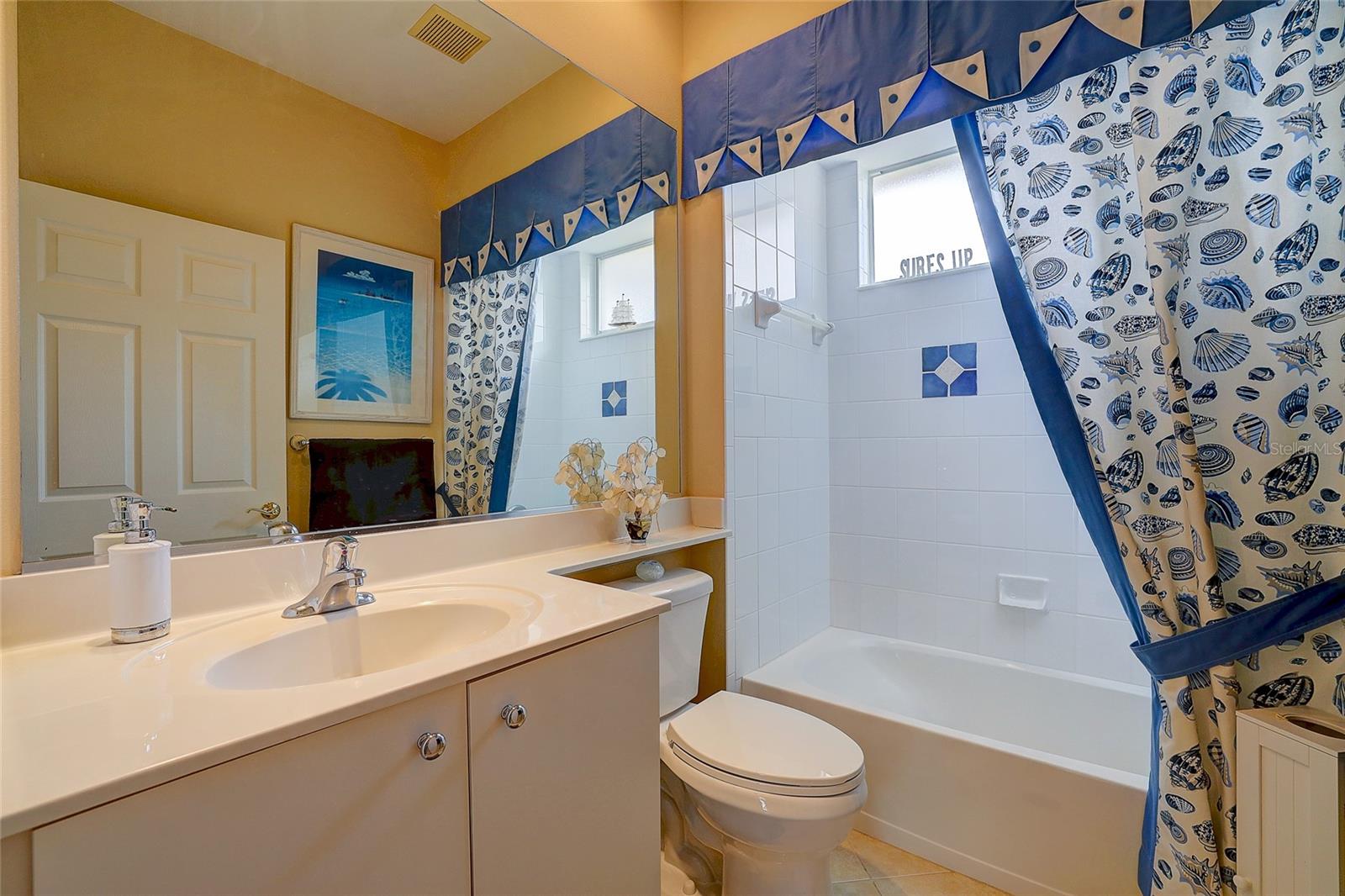 Bathroom with a white vanity, tiled tub-shower, and blue-and-white shell-print curtains.