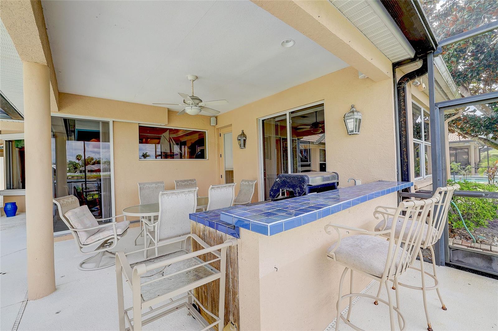 Covered patio with a blue-tiled bar, dining chairs, ceiling fan, and screened pool view.
