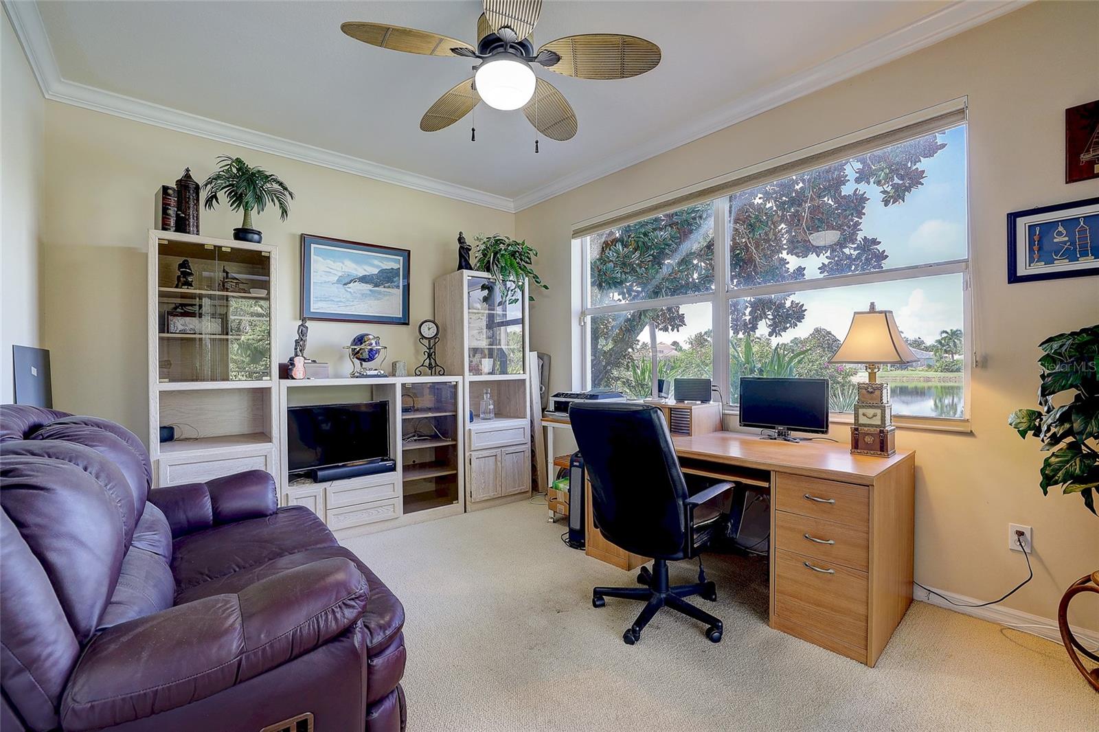 Furnished home office with a desk and purple sofa beside a large window overlooking a pond.