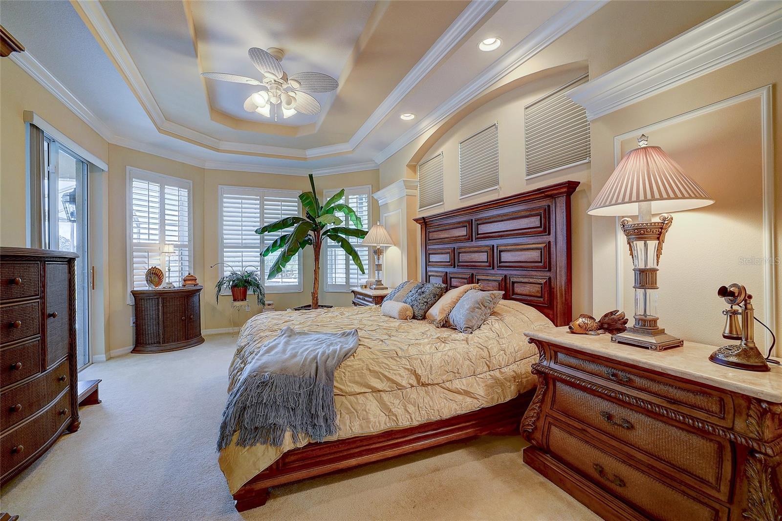 Furnished bedroom with a carved wood bed, plantation shutters, and a tray ceiling with ceiling fan.