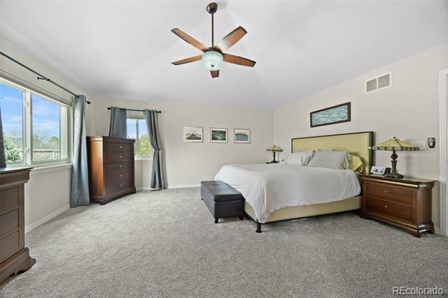 Furnished bedroom with a large bed, gray carpet, tall curtained windows, and a ceiling fan.
