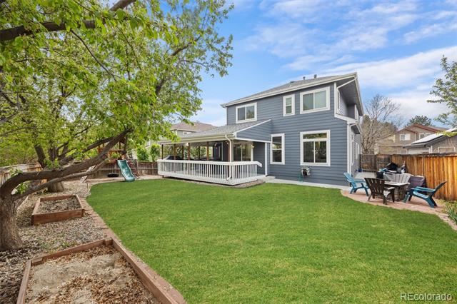 Two-story blue-gray house with a covered deck, broad lawn, mature tree, and outdoor dining area.