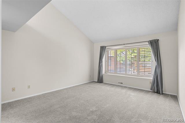 Vacant carpeted room with a high sloped ceiling and a wide shuttered window framed by gray curtains.