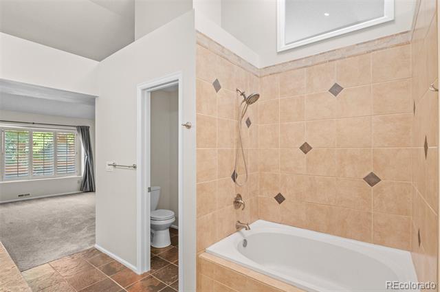 Bathroom with a white bathtub, tan diamond-accented tile, and an open doorway to a toilet and carpeted room.