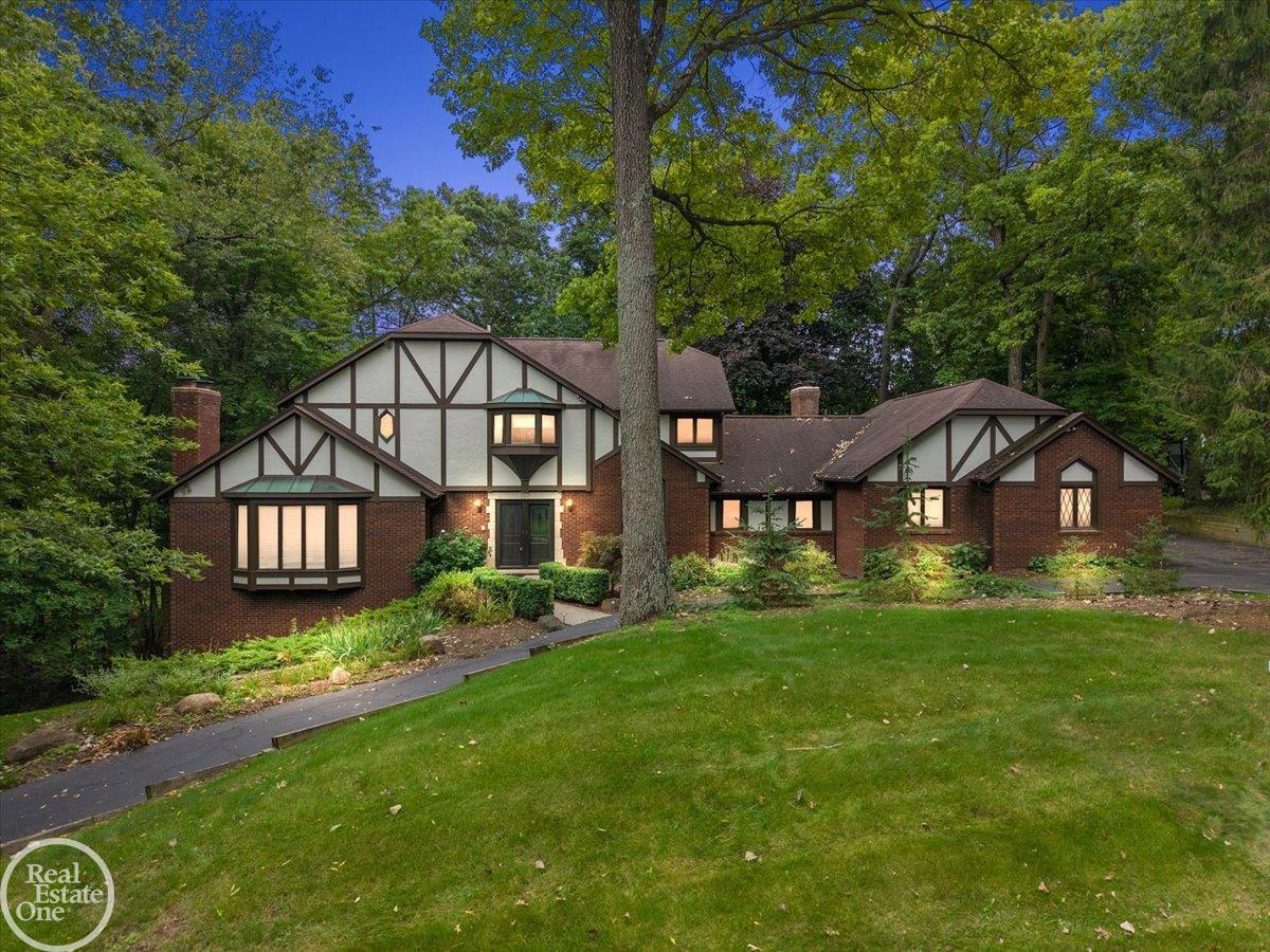 Tudor-style brick home with a wooded setting, broad lawn, and warmly lit windows.