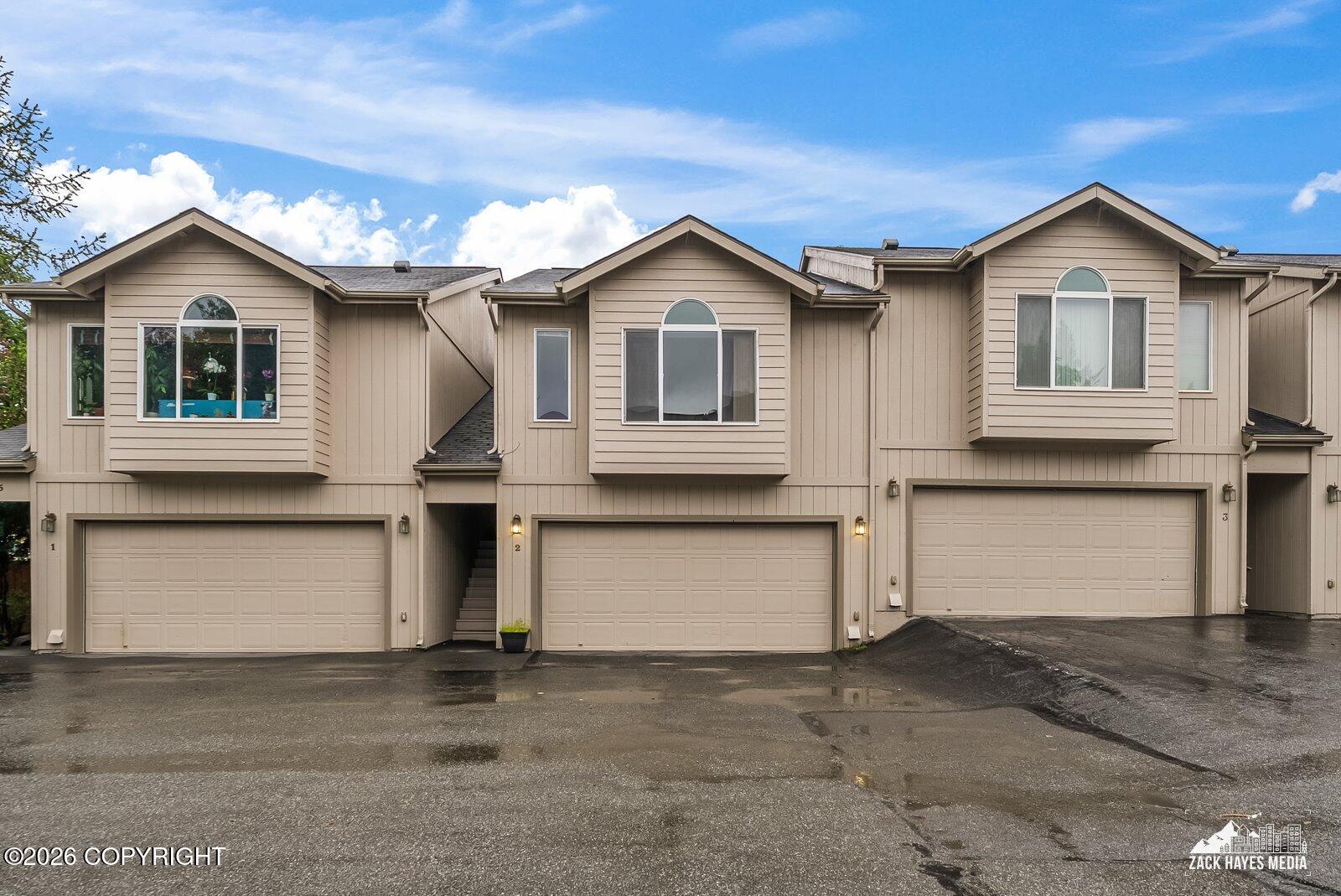 Three adjoining beige townhomes with peaked gables and individual garages along a wet driveway.