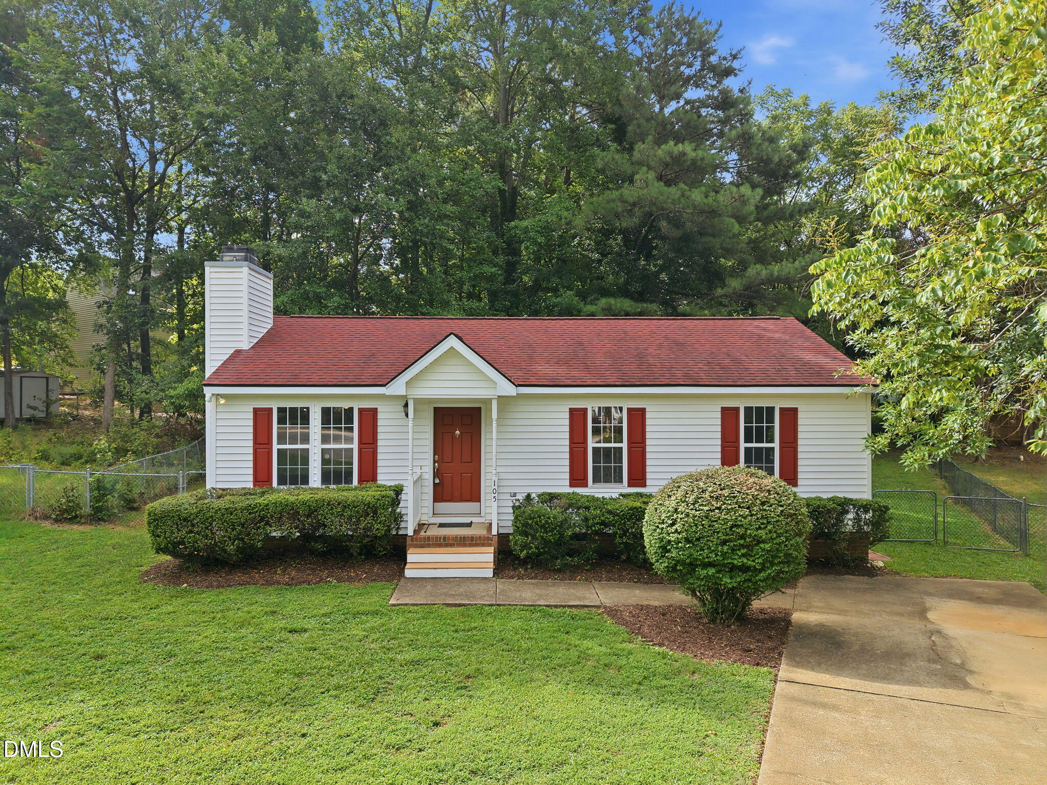 White single-story house with a red roof and shutters, trimmed shrubs, and a concrete driveway.