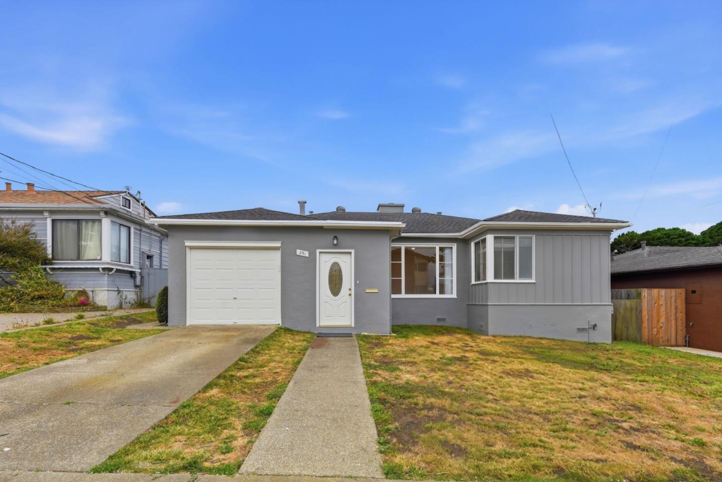 Single-story gray house with a white garage door, wide driveway, and patchy front lawn.