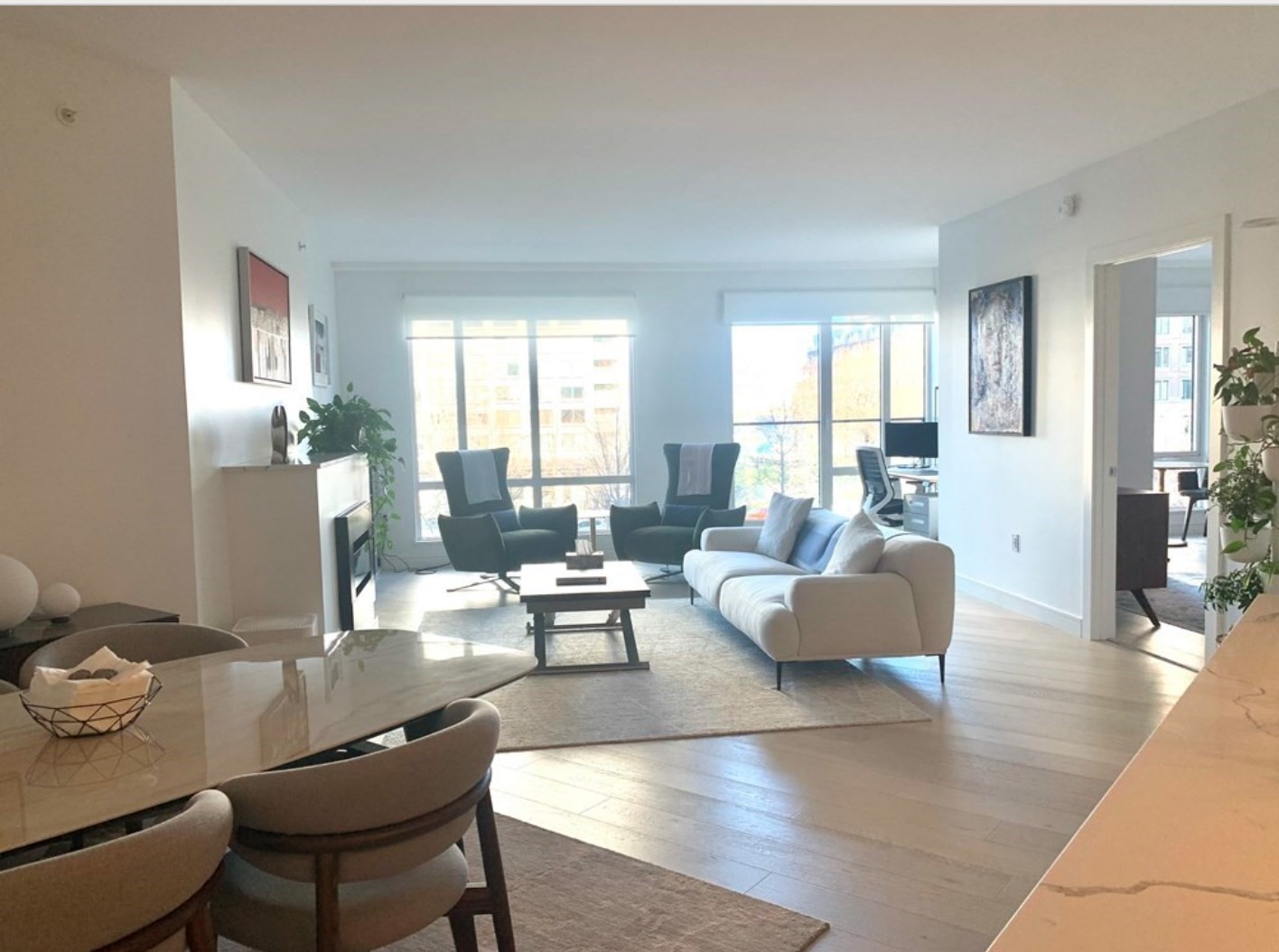 Bright condo living room with large windows, white sofa, and dark armchairs beside the dining area.