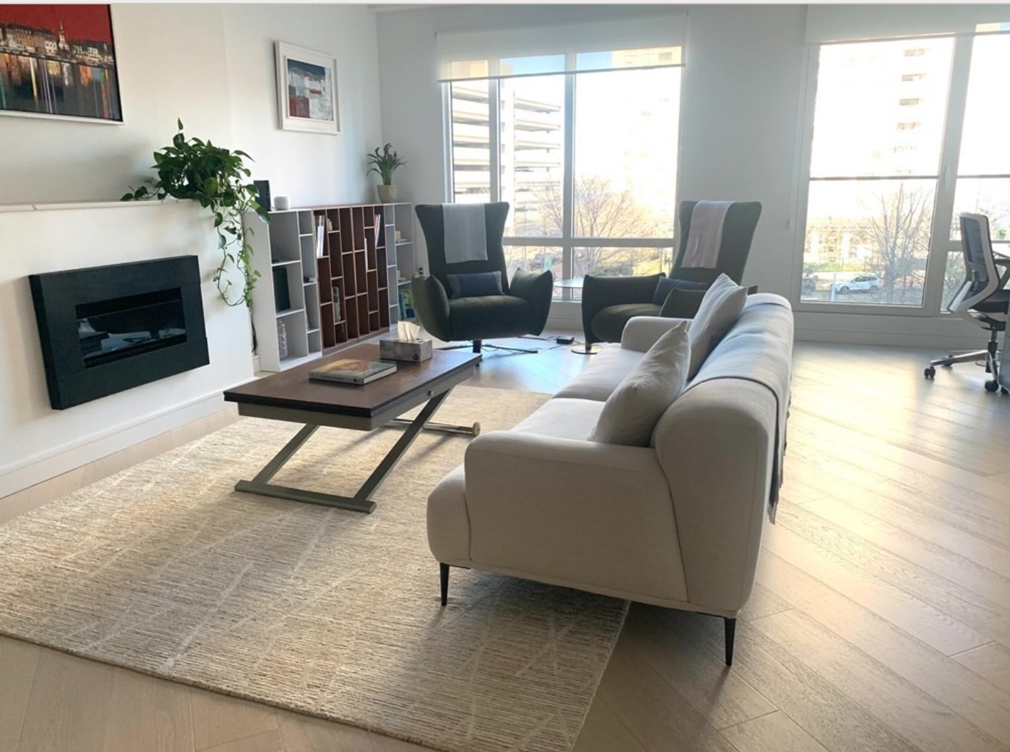 Modern living room with a light sofa, wall-mounted fireplace, and large windows overlooking nearby buildings.