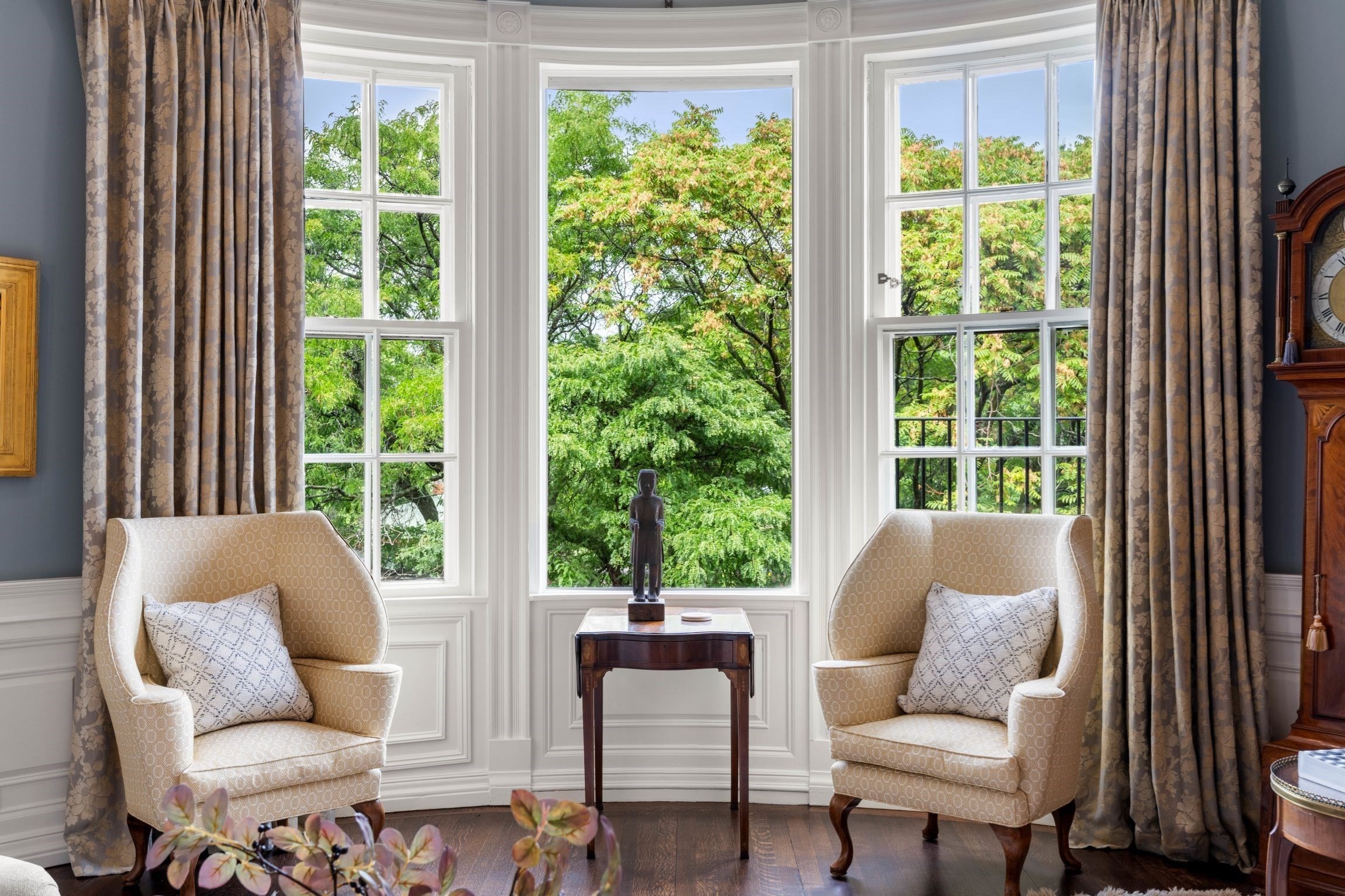 Living room with paired armchairs, a small table, and tall bay windows overlooking leafy trees.
