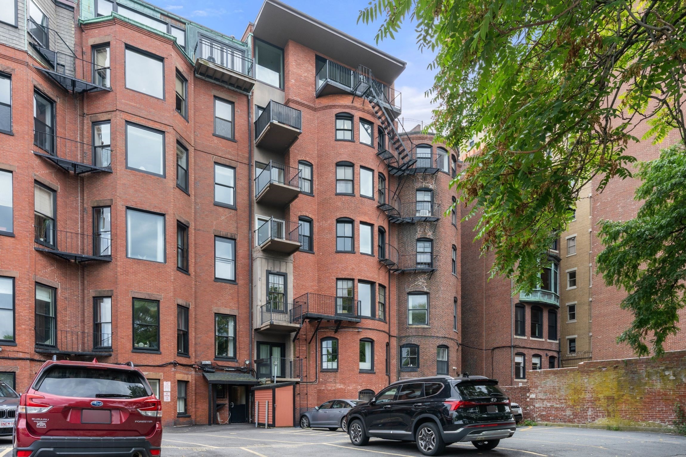 Red-brick apartment building with balconies and fire escapes beside a small parking area.