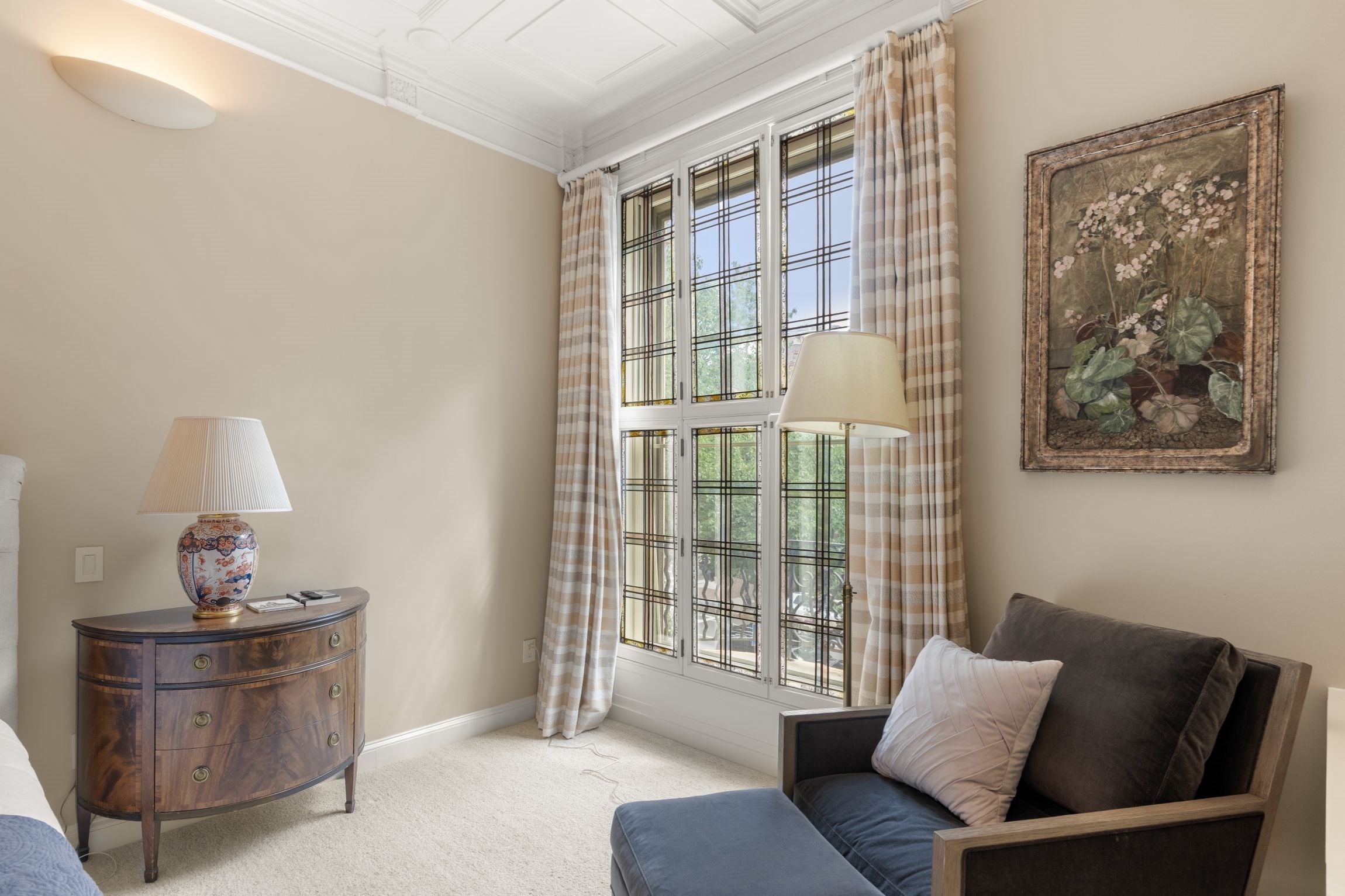 Furnished bedroom with tall divided-light windows, a curved wooden dresser, and a dark upholstered armchair.