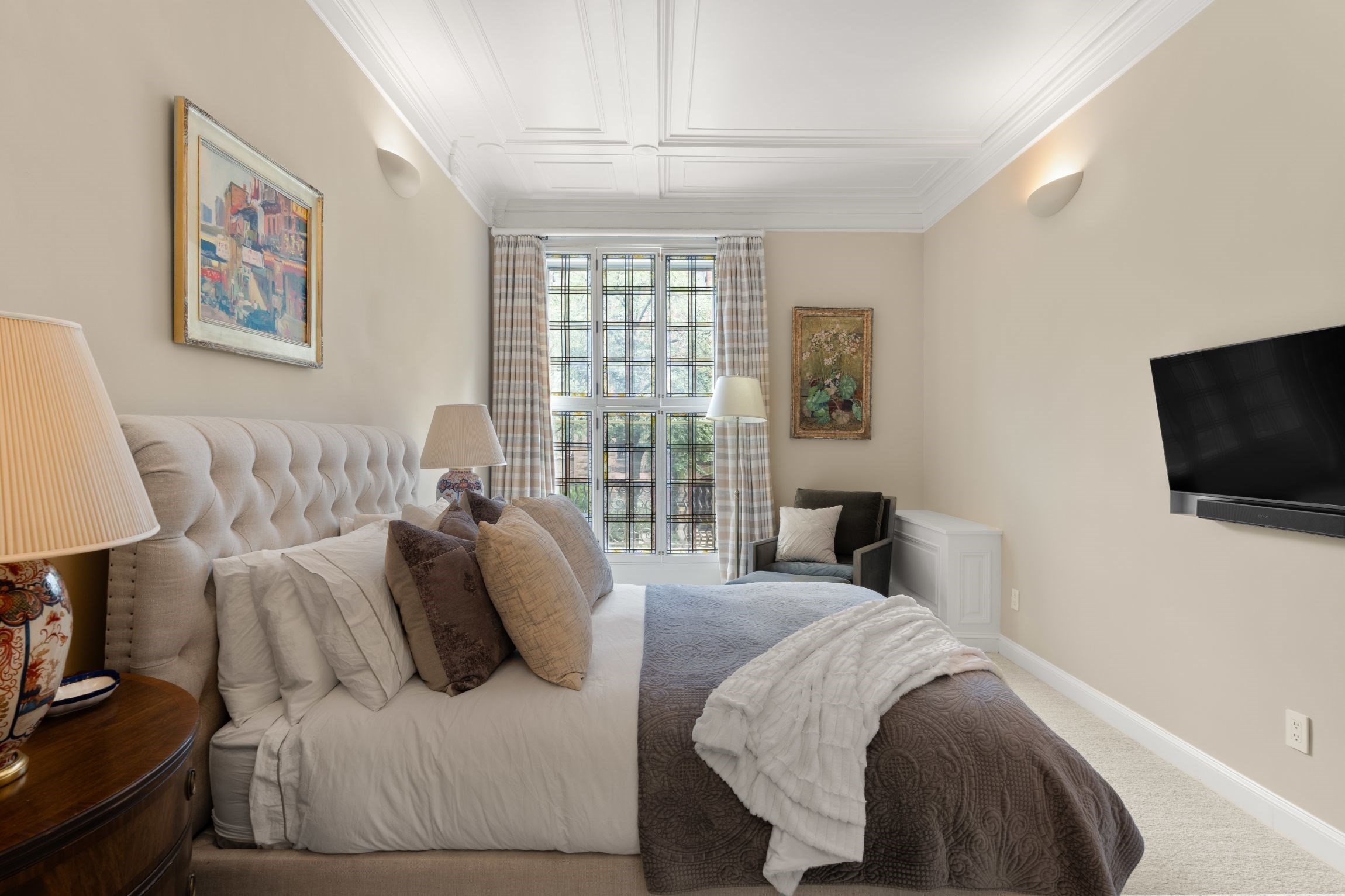 Furnished bedroom with a large upholstered bed beneath a decorative ceiling and tall multi-pane window.