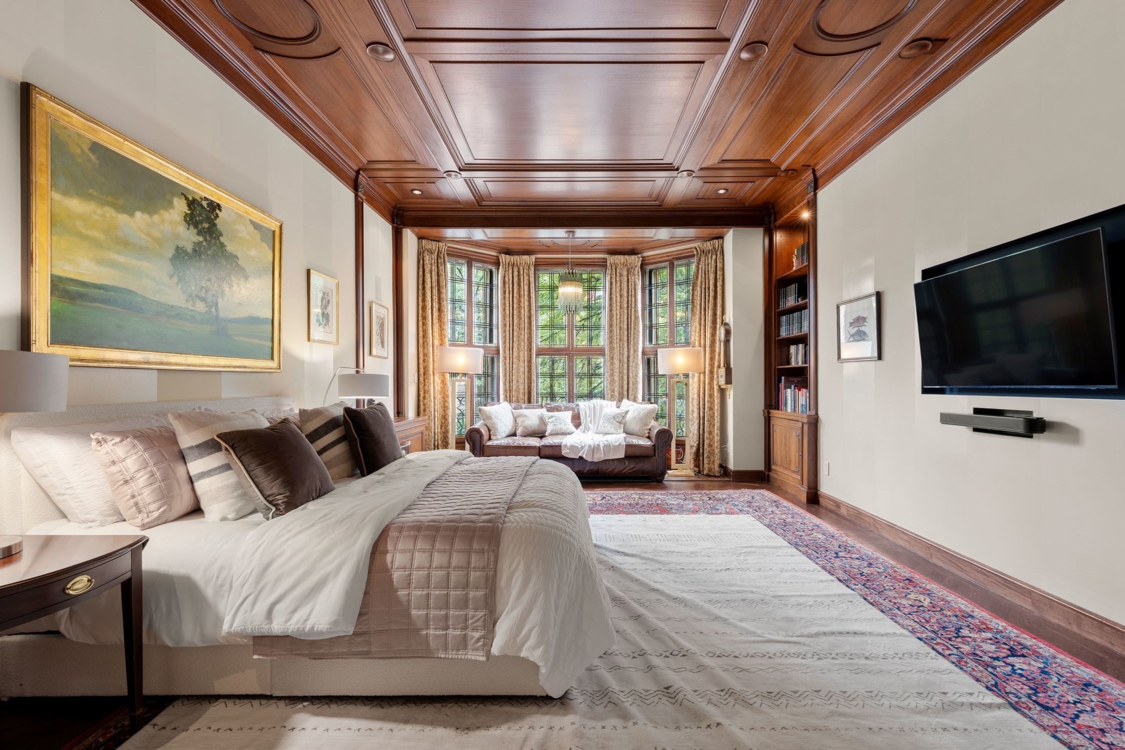 Bedroom with a richly paneled wood ceiling, large bed, and window-side sitting area.