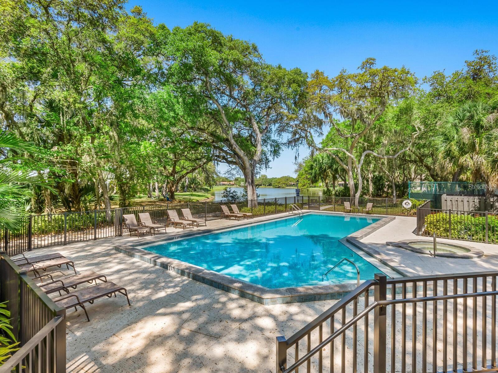 Community pool and spa beside a tree-lined lake, with lounge chairs on the sunny deck.