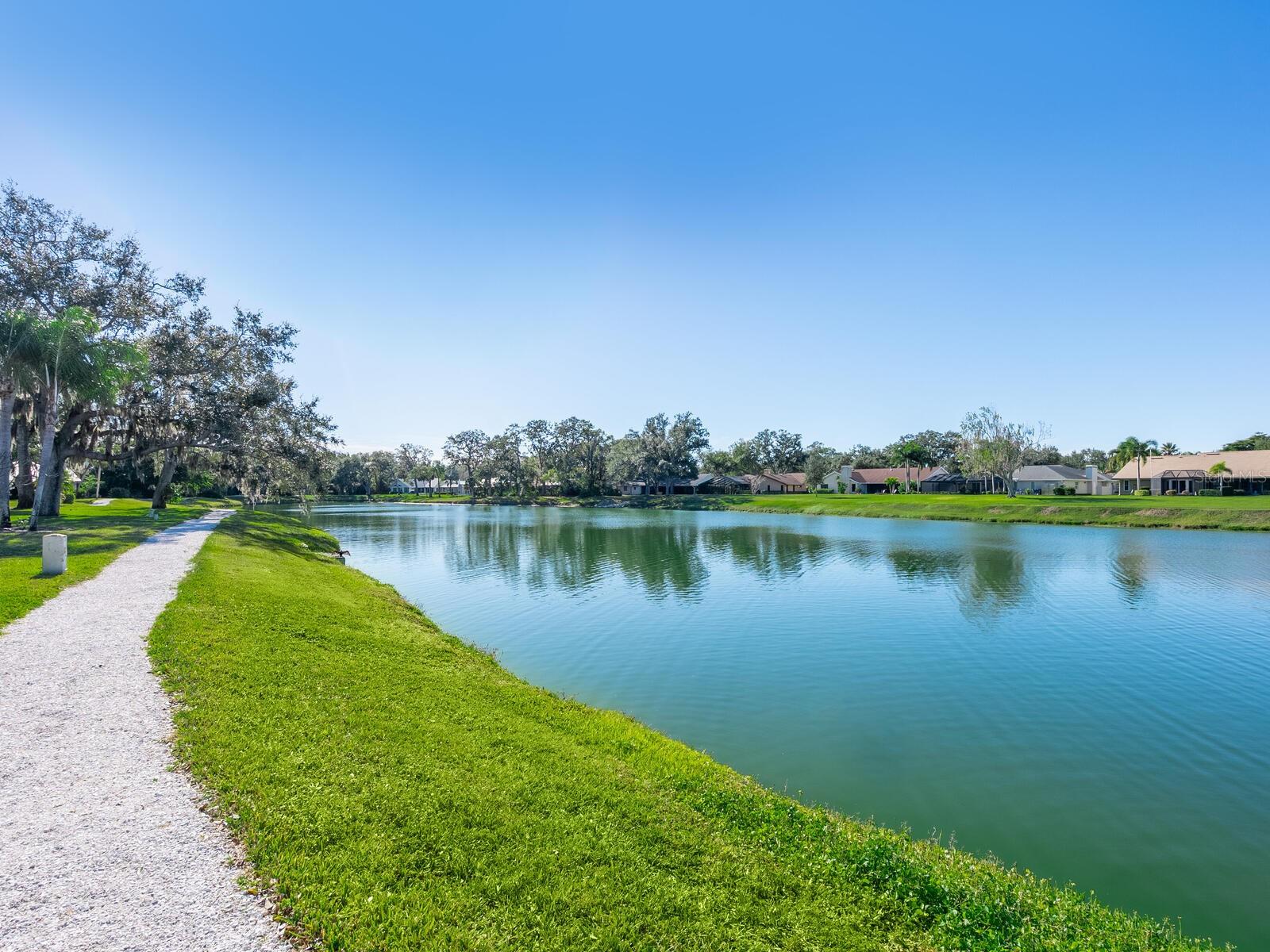 Community pond with a gravel walking path, grassy banks, and homes across the water.