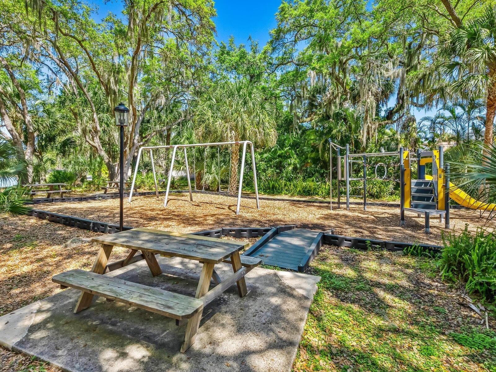Community playground with swings, climbing equipment, and a picnic table beneath mature trees.