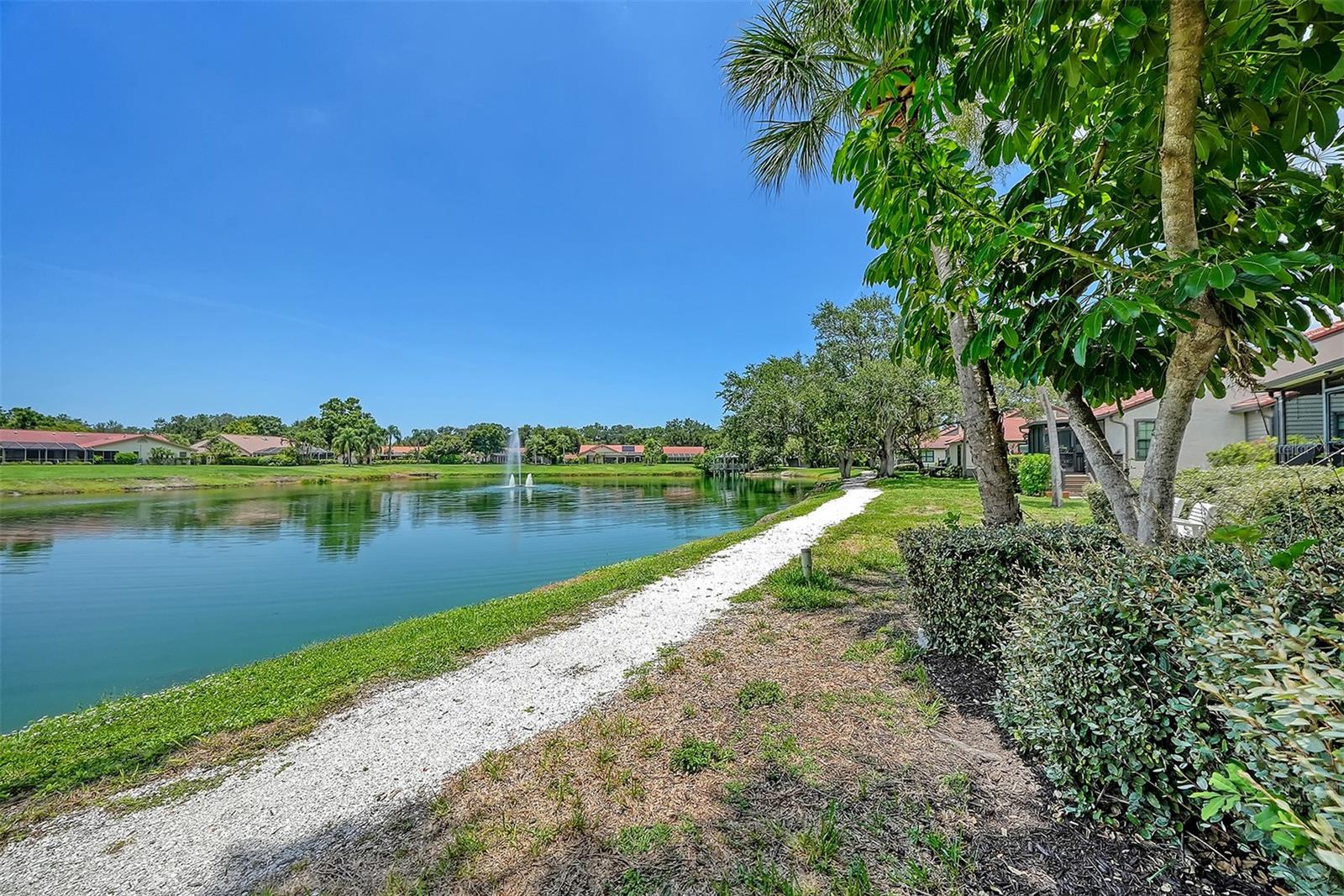 Pond and fountain beside a gravel path, with palms and residences along the shoreline.