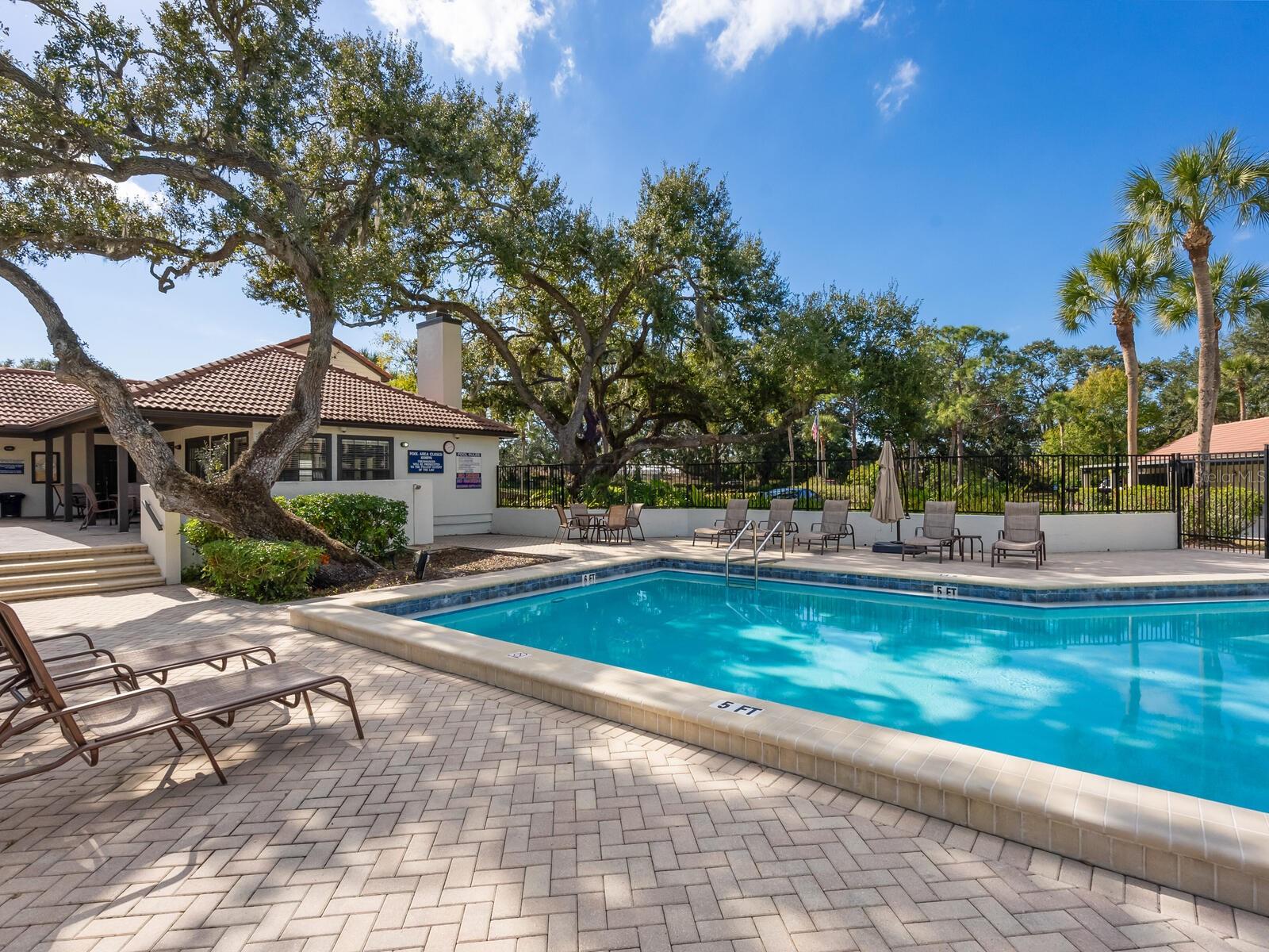 Community swimming pool with lounge chairs, a paved deck, and mature oak and palm trees.