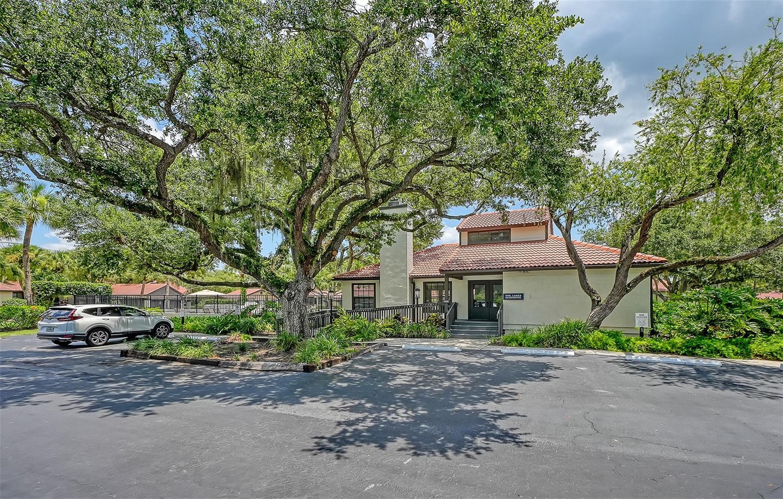Condominium building beneath sprawling oak trees, with a red tile roof and landscaped entrance.