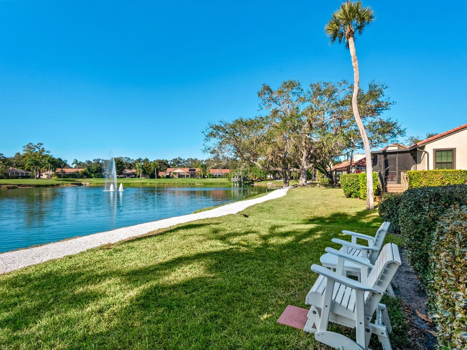 Residential pond with fountains, a curving gravel shore, and white Adirondack chairs on the lawn.