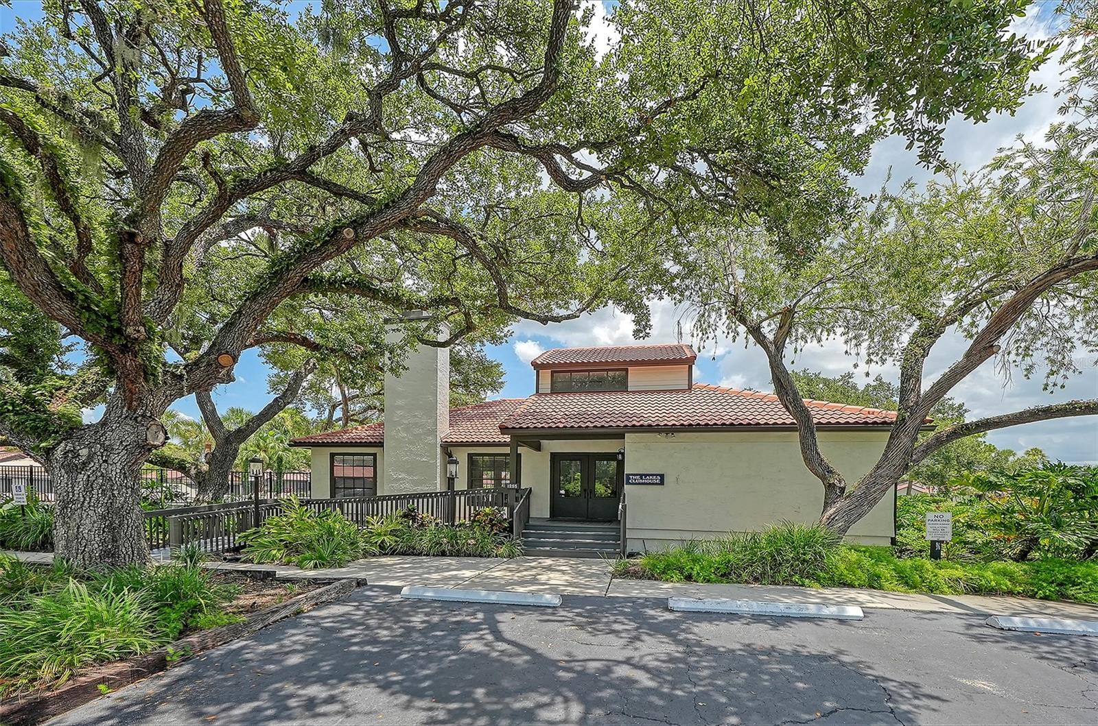 Clubhouse beneath mature trees, with a red tile roof and landscaped entrance.