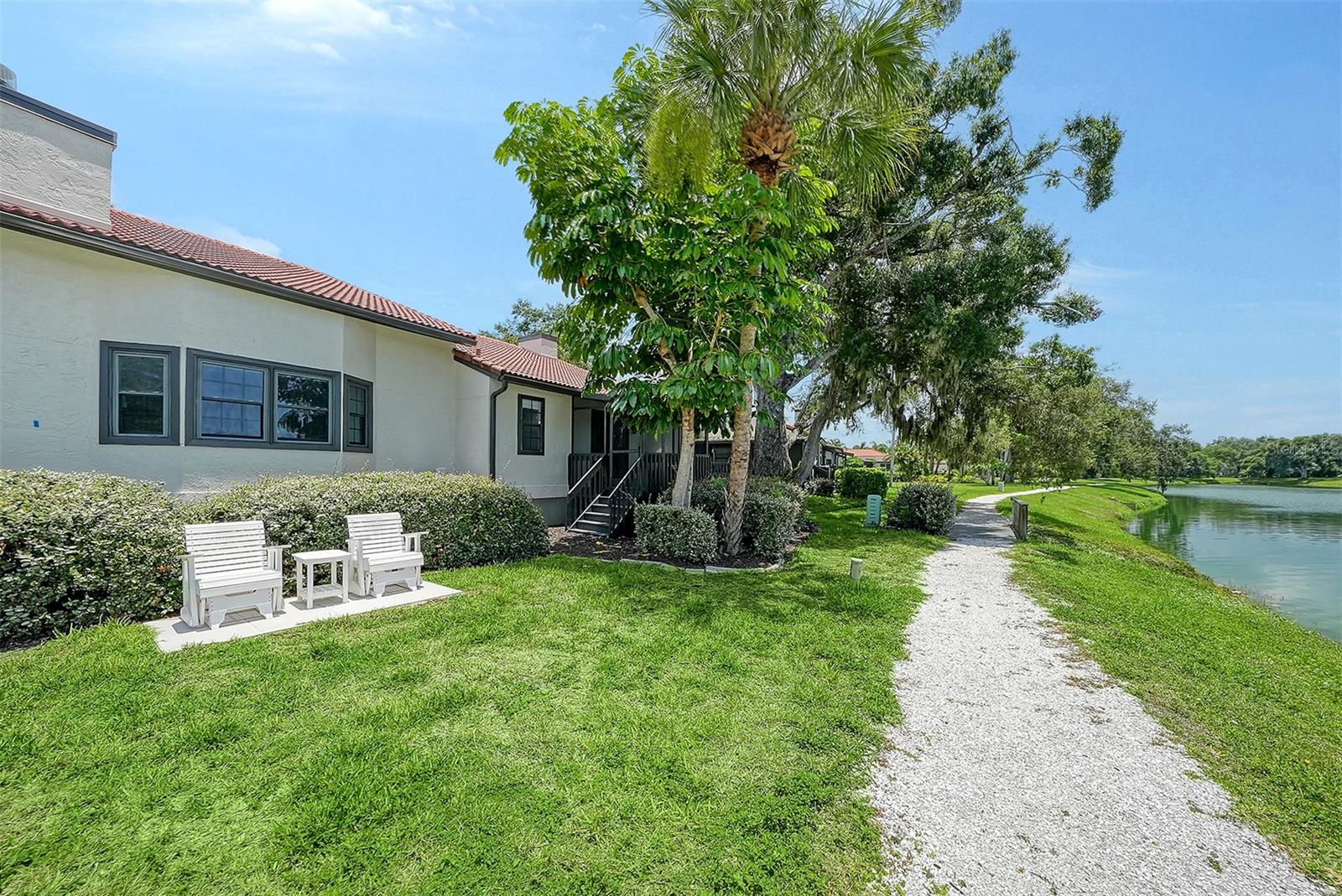 Home beside a pond, with a gravel path, palm trees, and white outdoor chairs.