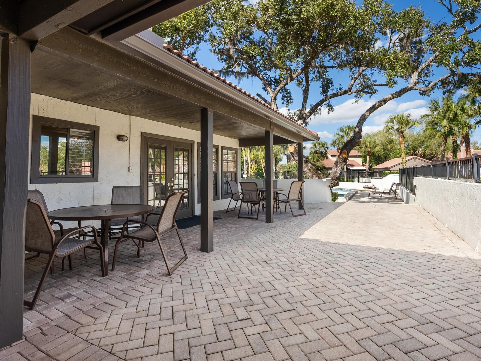 Covered patio with outdoor dining furniture and brick paving beside a backyard pool.
