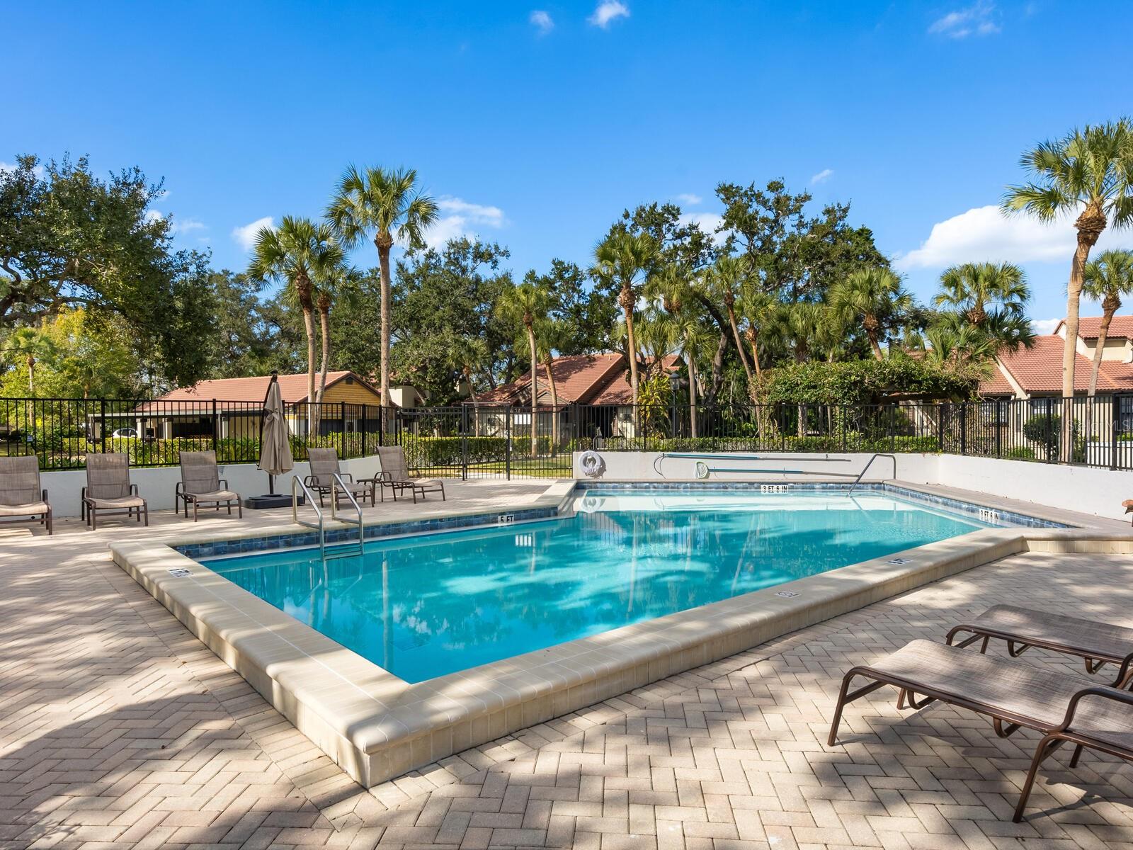 Outdoor pool with a broad paver deck, lounge chairs, palm trees, and a fenced perimeter.