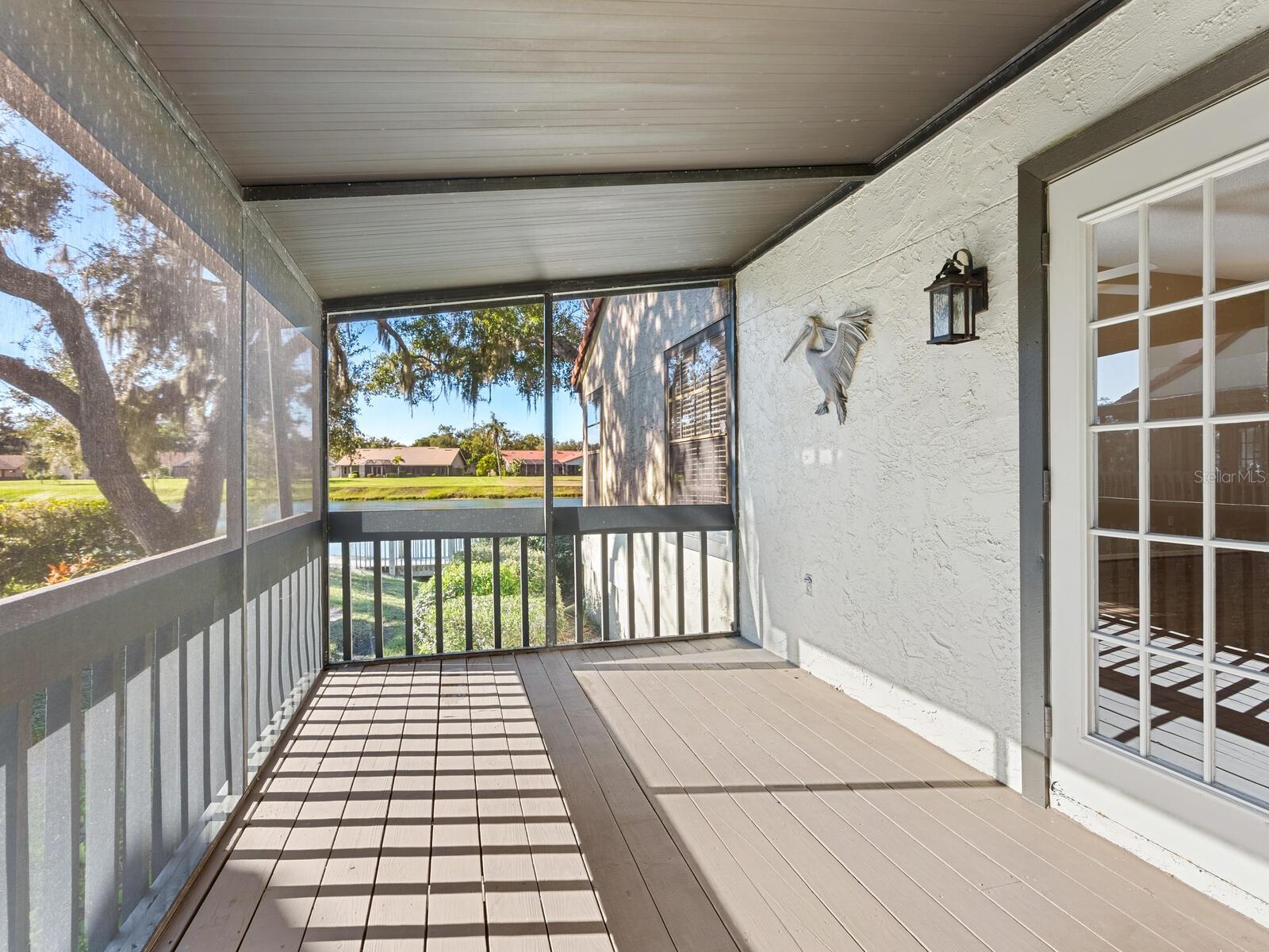 Screened porch overlooking a pond, with tan plank flooring and sunlight casting shadows across the floor.