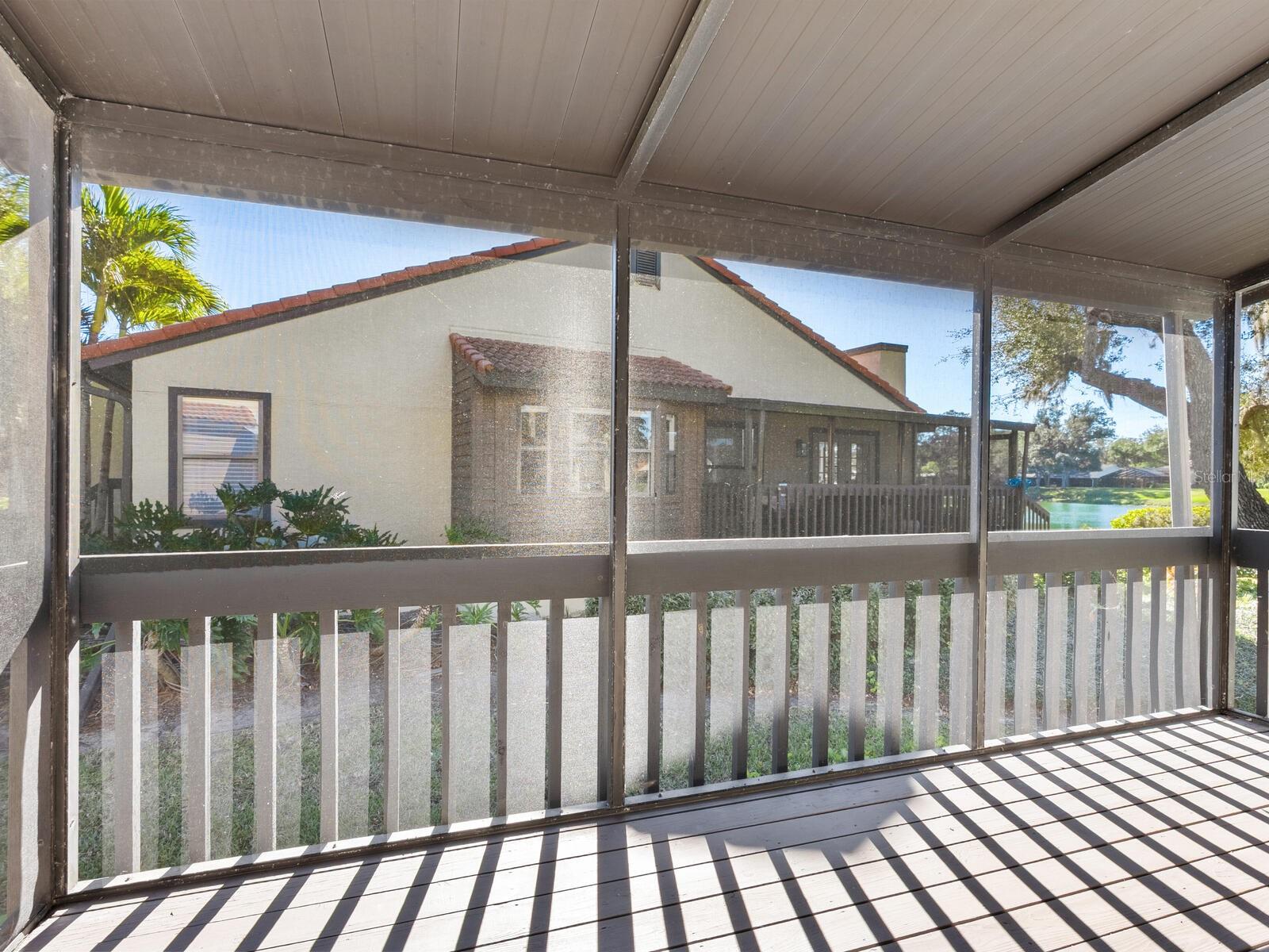 Screened porch overlooking a neighboring home and lake, with sunlight casting striped shadows across the deck.