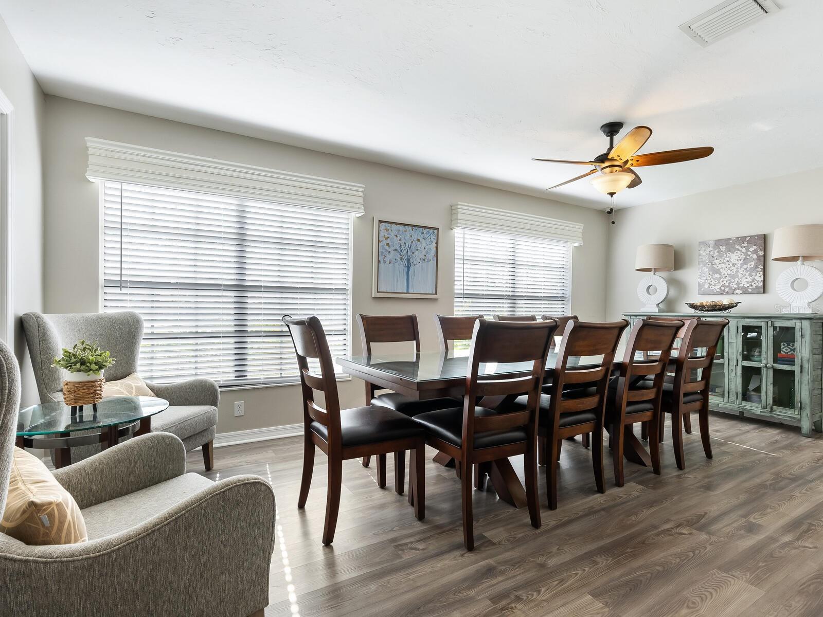 Dining room with a long dark wood table, gray seating, and wide windows with white blinds.