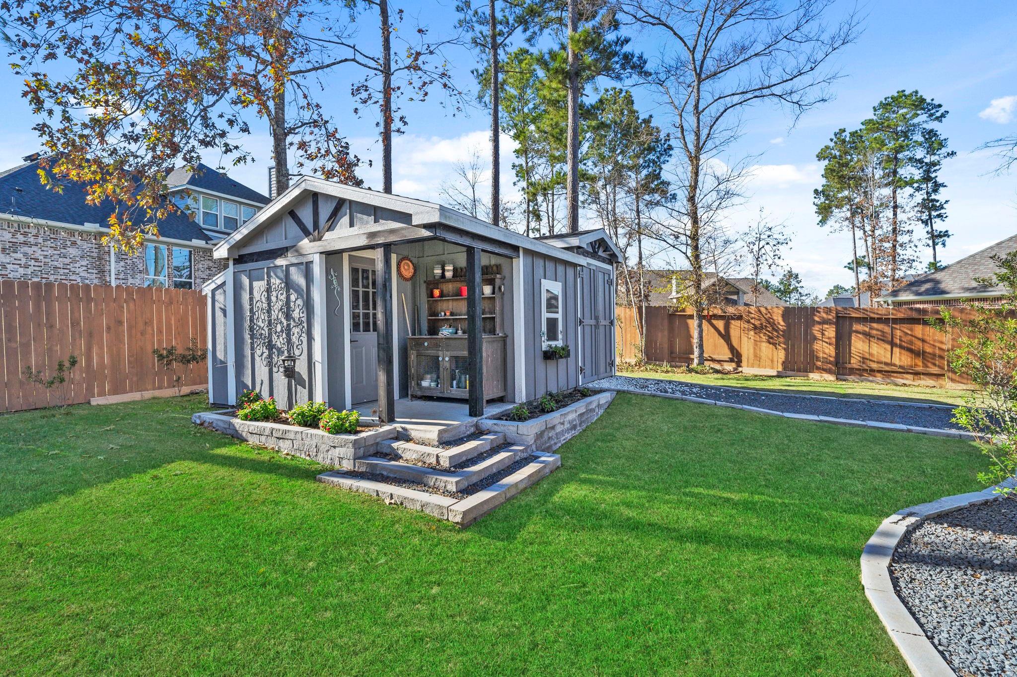Gray garden shed with a covered porch, stone steps, and flower beds in a fenced backyard.