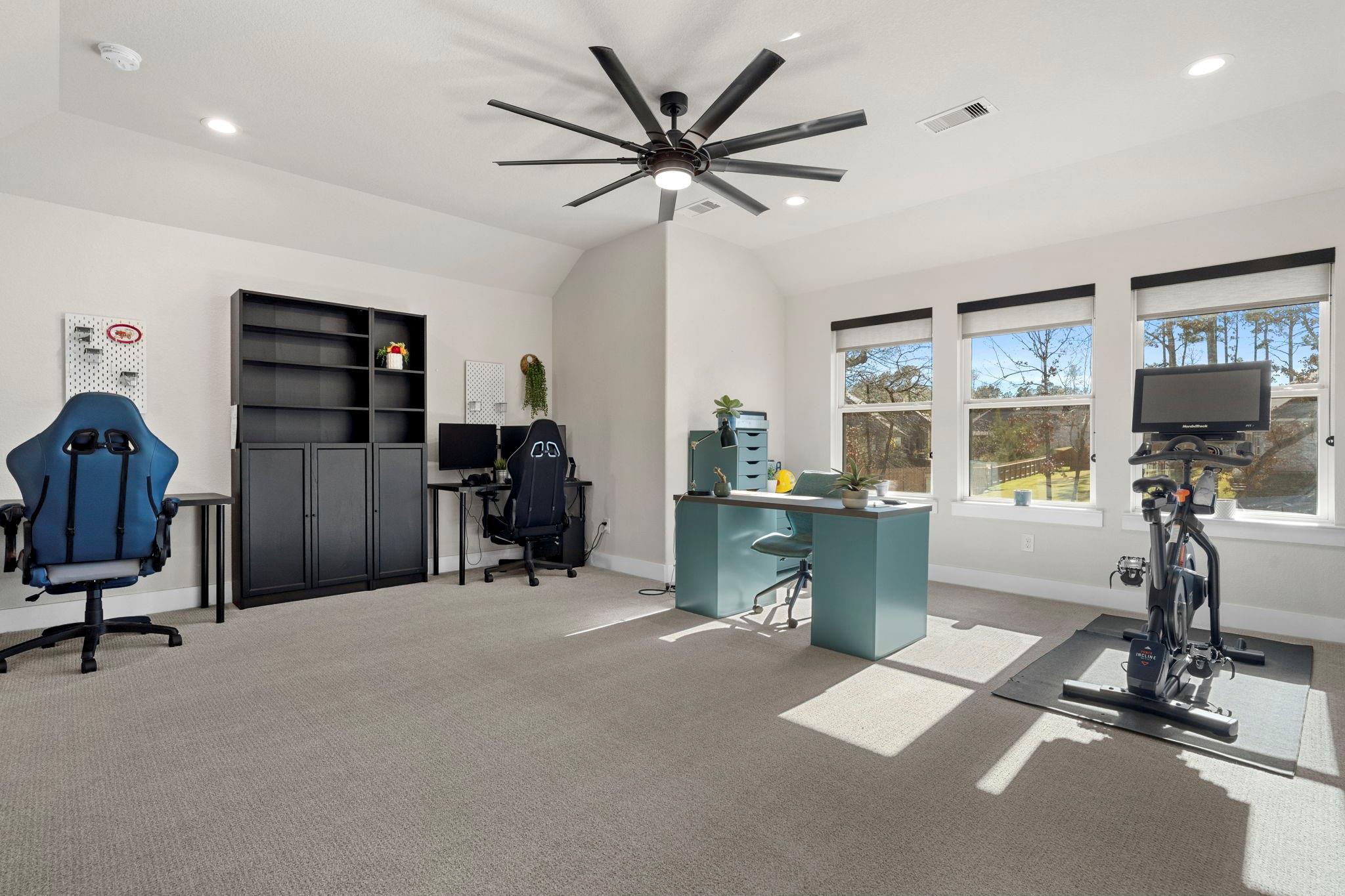 Home office and workout room with a teal desk, gaming chairs, and an exercise bike by large windows.