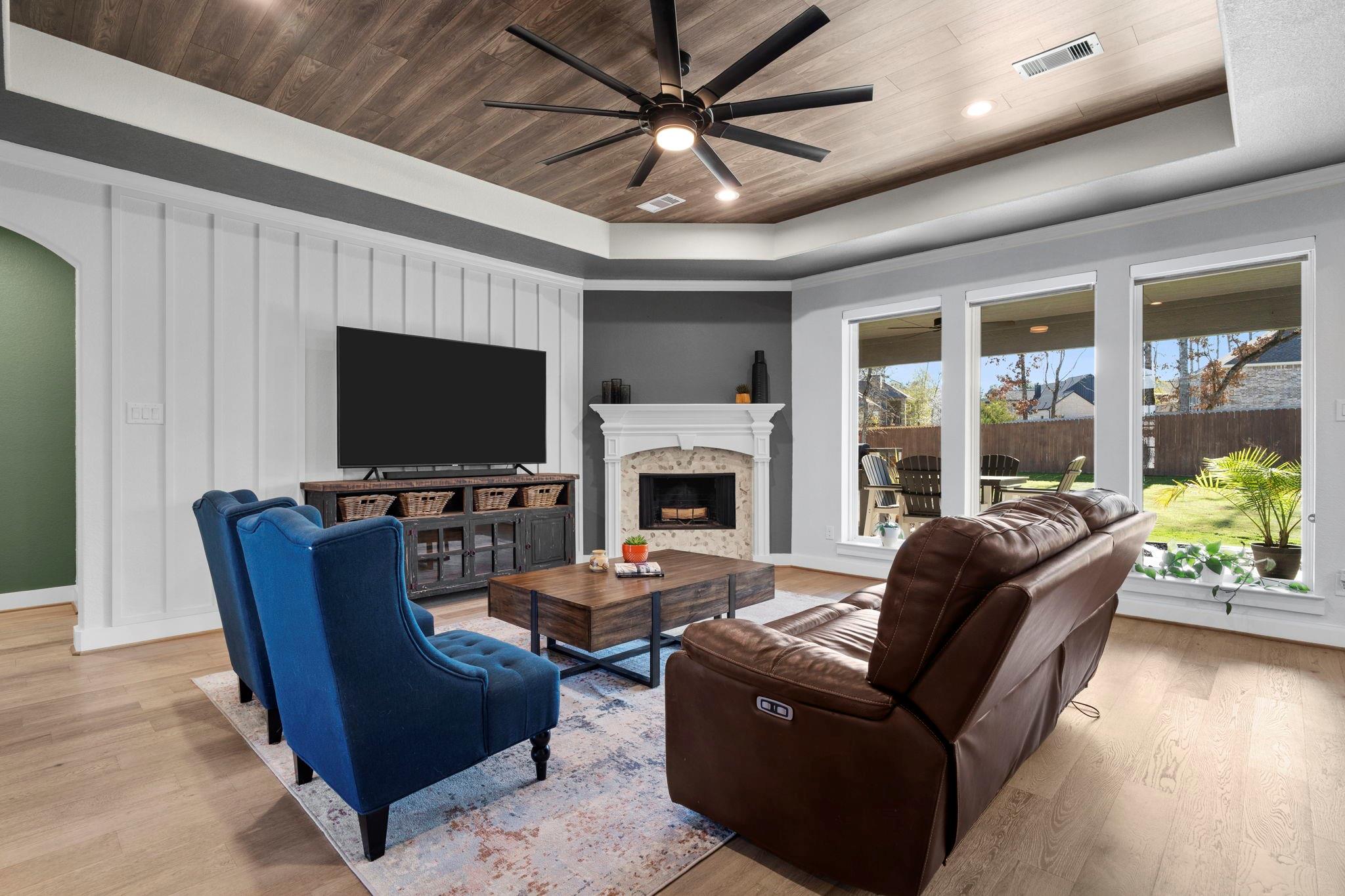 Living room with a stone fireplace, blue armchairs, leather sofa, and large windows overlooking the backyard.