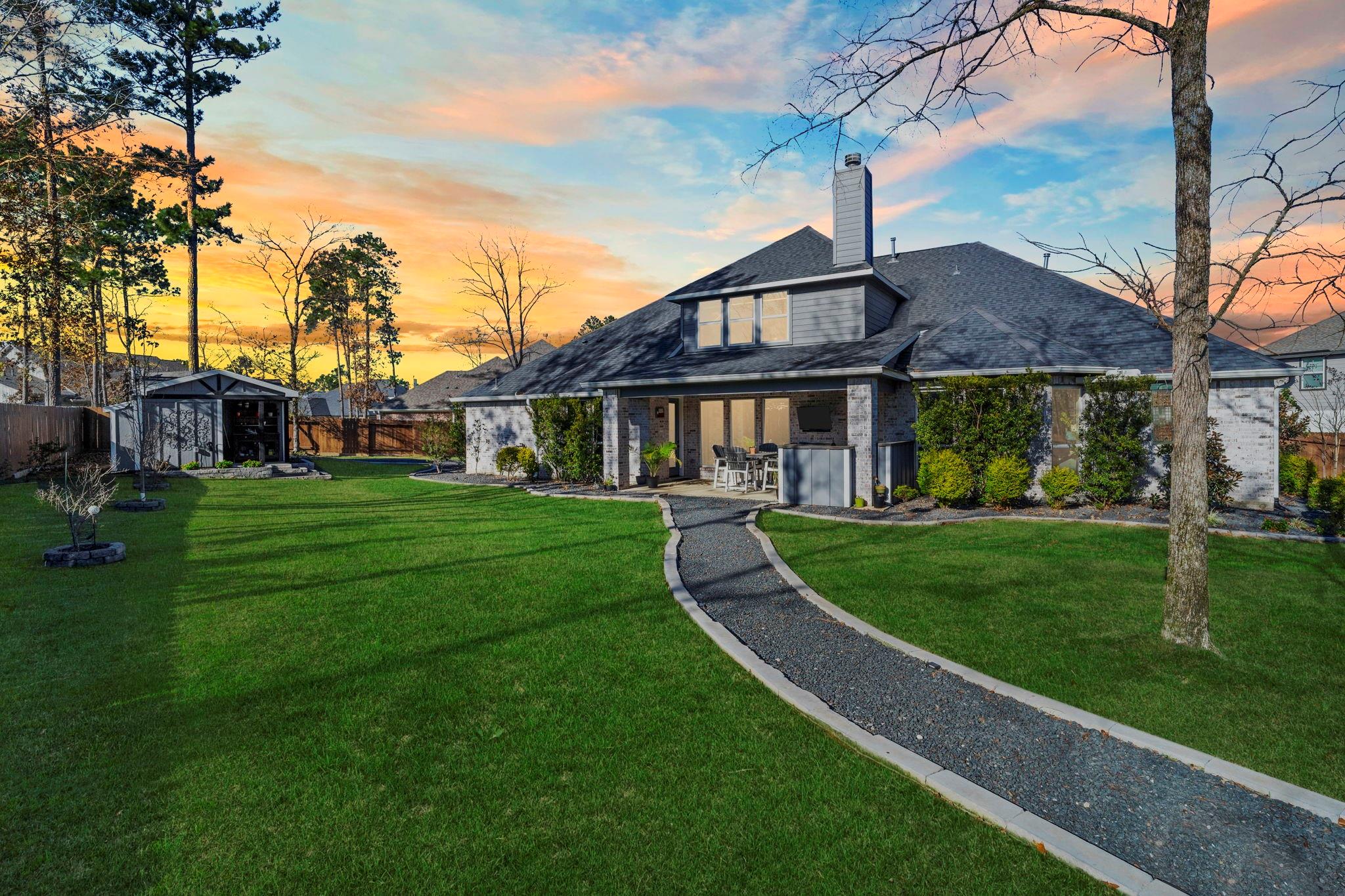 Rear of a two-story home with a covered patio, curved gravel path, and detached pavilion at sunset.
