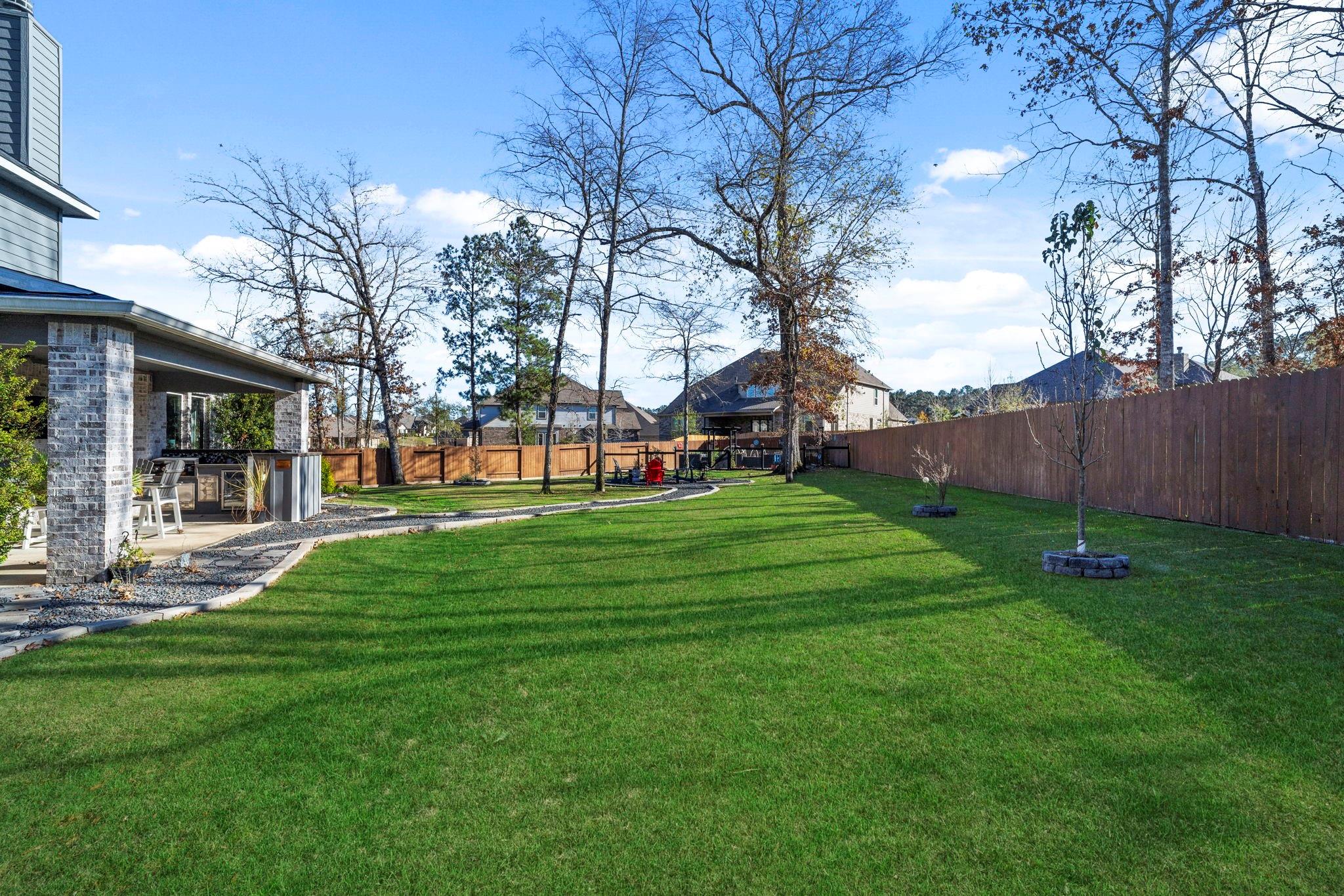 Fenced backyard with a large lawn, mature trees, and a covered patio with a built-in grill.