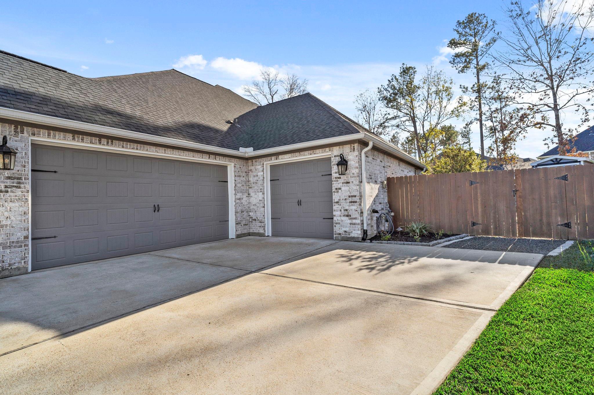 Brick home garage with two gray doors, a wide concrete driveway, and a tall wooden privacy fence.
