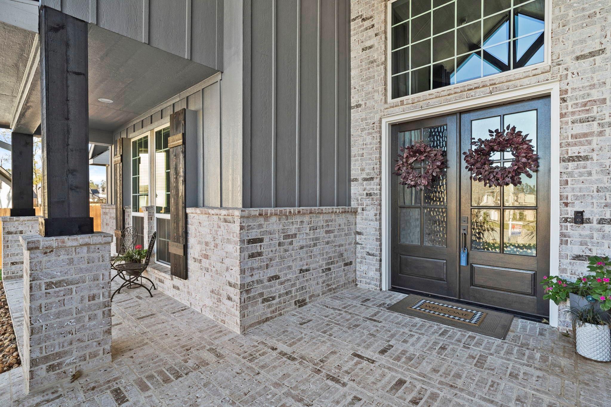 Home’s covered front porch with brick paving, dark double doors, wreaths, and a small seating area.