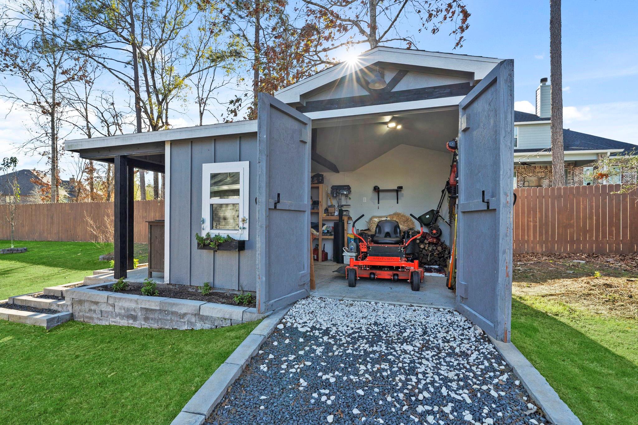 Gray garden shed with open doors, a riding mower inside, and a gravel path across the lawn.