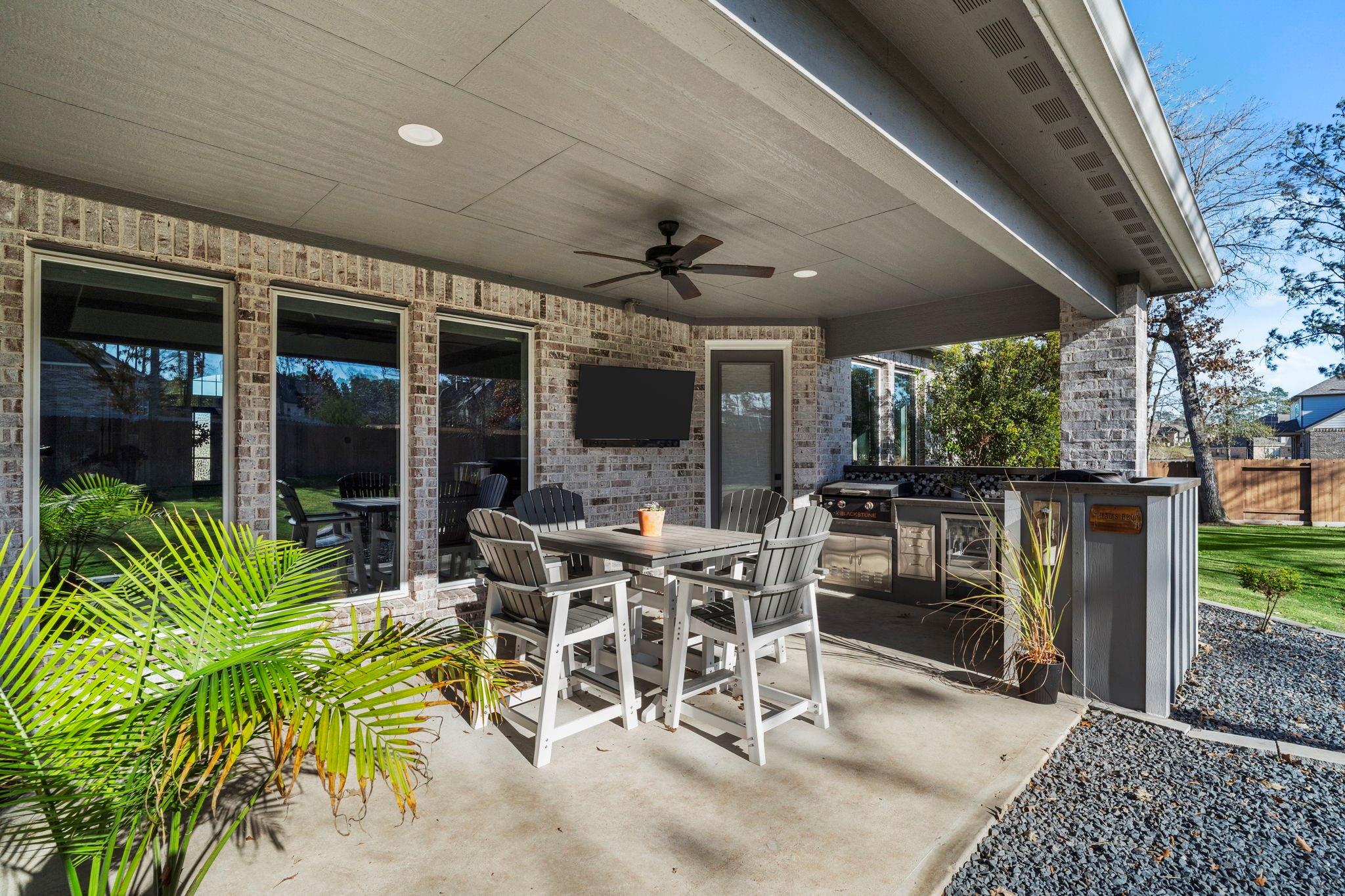 Covered brick patio with outdoor dining set, ceiling fan, television, and built-in grill.