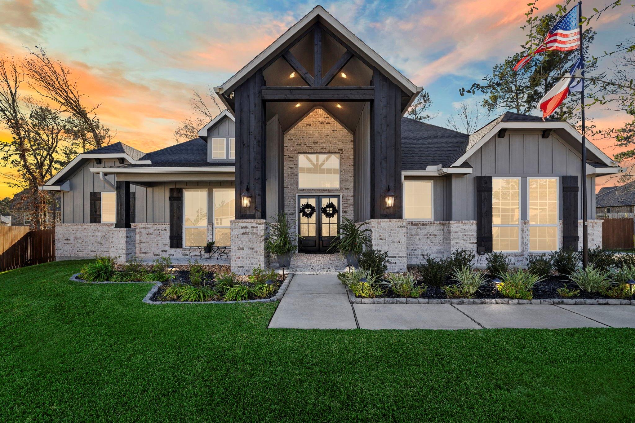 Modern farmhouse exterior with a tall timber entry gable, landscaped lawn, and American flag at dusk.