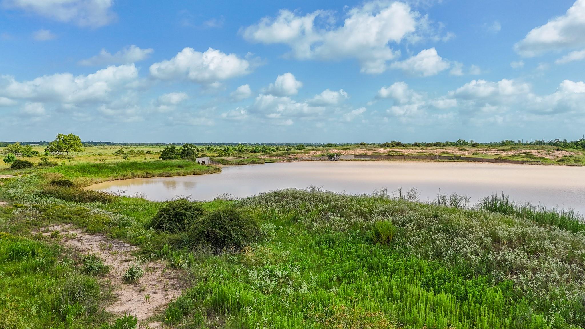 Open grassy land surrounds a large pond, with a drainage structure across the water.